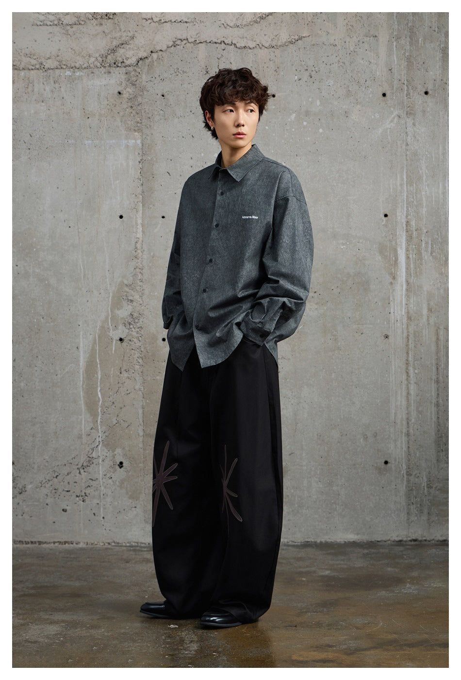 Model wearing a black gray washed-look button-up shirt over wide black pants, standing against a concrete wall