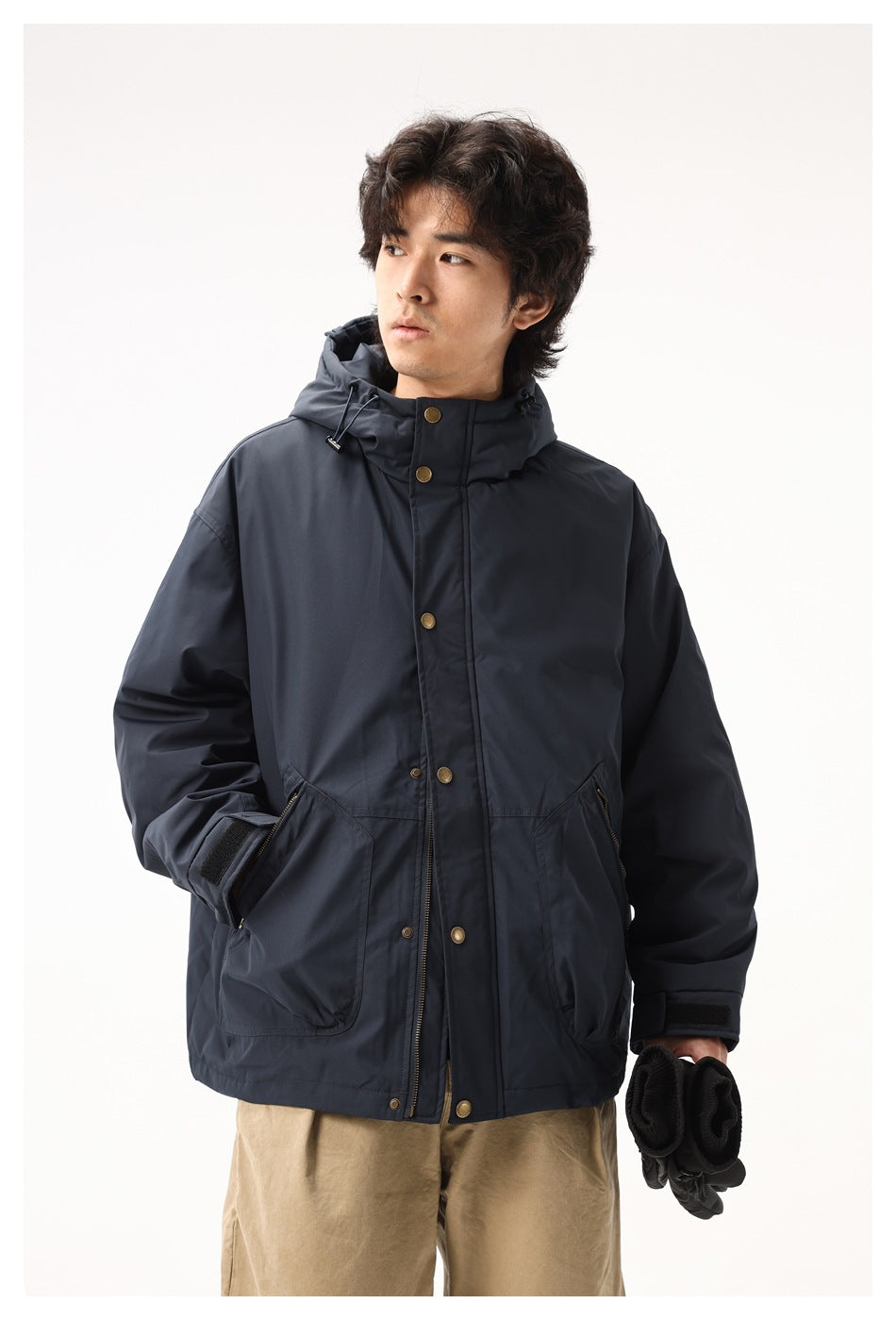 Model wearing a dark navy hooded jacket with brass-tone snaps and large pockets, holding black gloves.