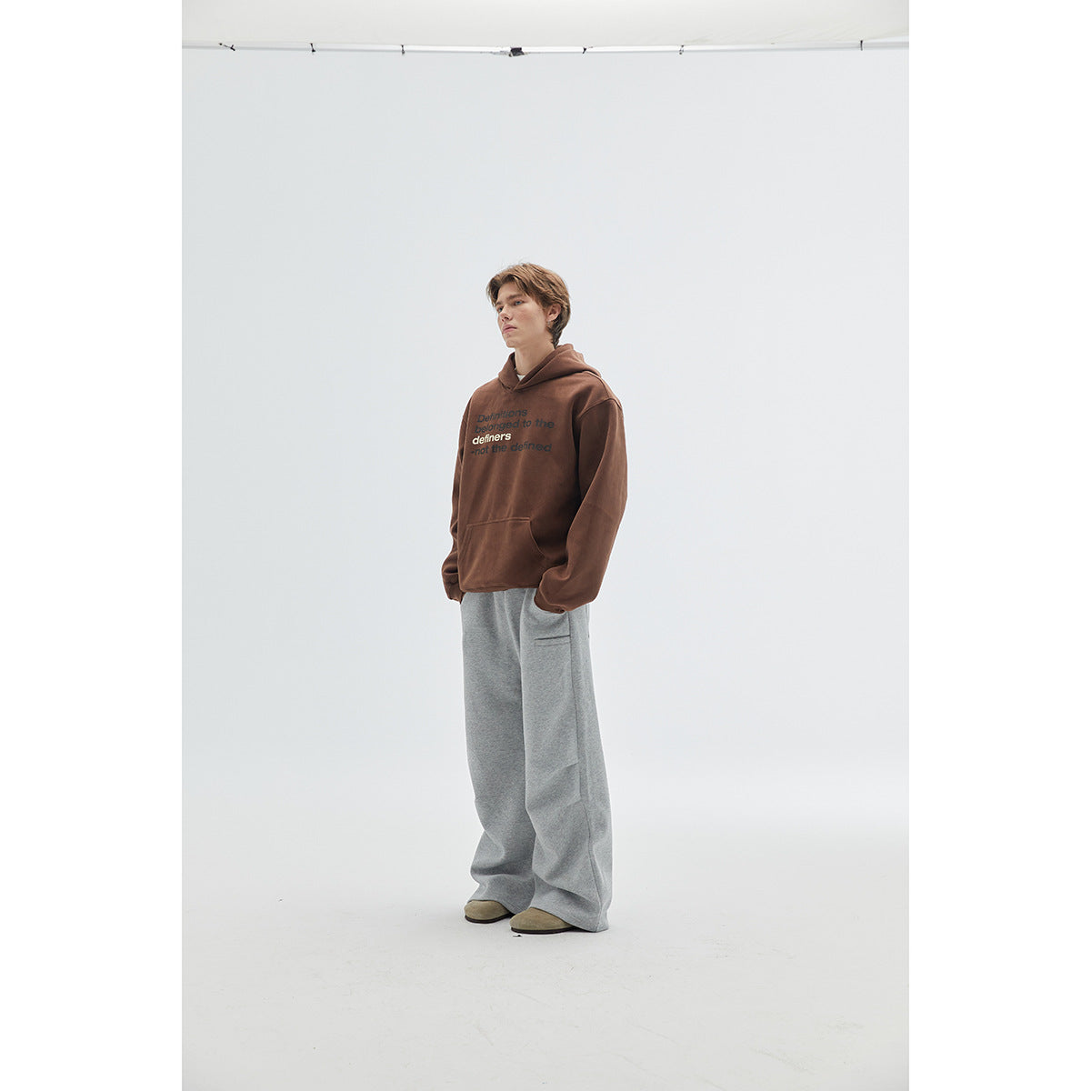 Model in a brown text-print hoodie and wide gray pants, posed at a three-quarter angle with hands in pockets.