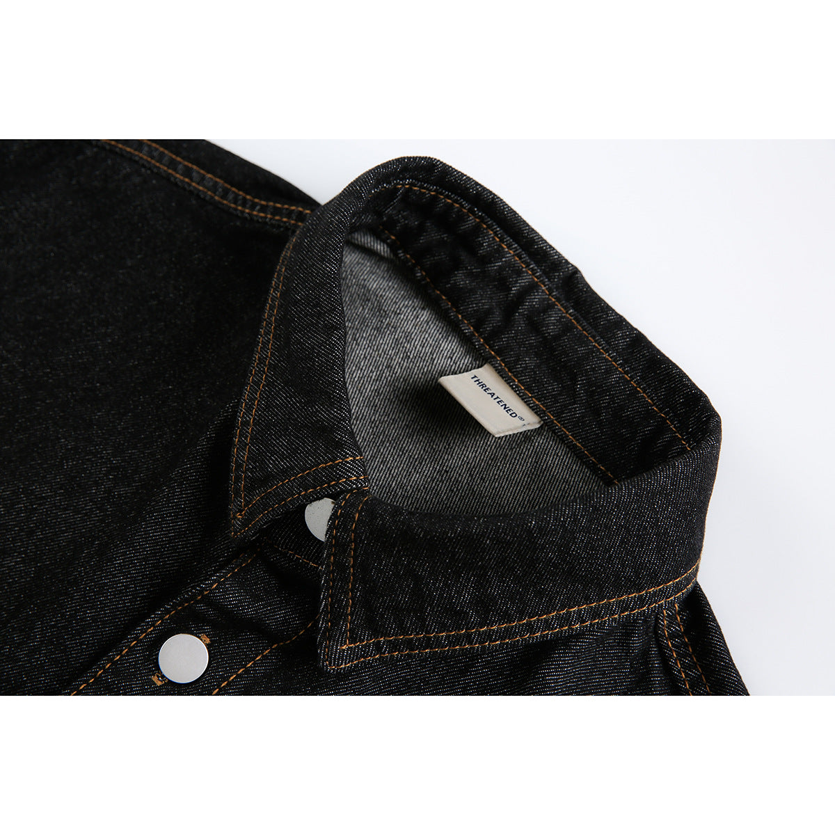 Close-up of a black denim shirt collar, inner label, and silver snap with orange stitching.