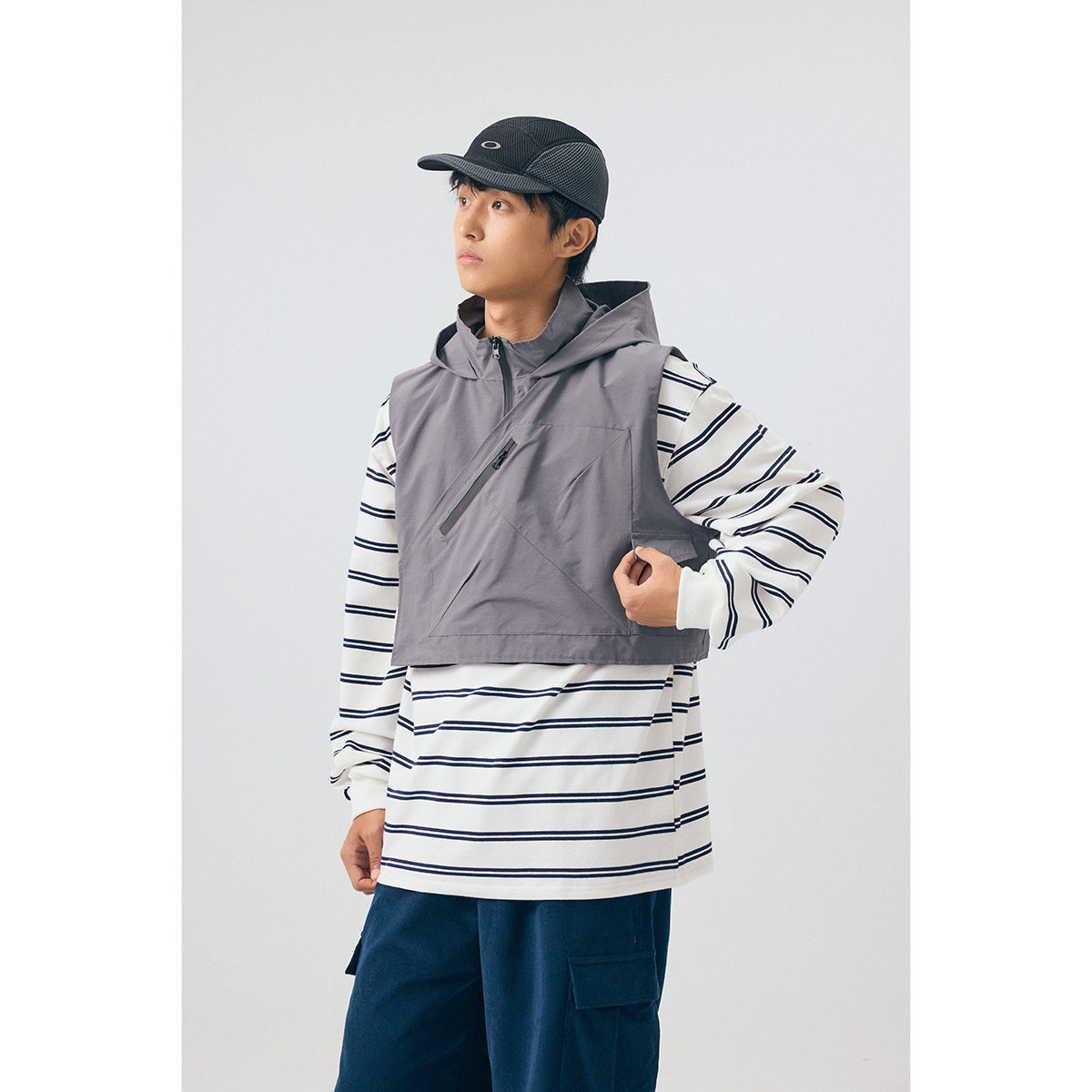 Model wearing a gray cropped hooded vest with a diagonal zip pocket over a white and navy striped shirt.
