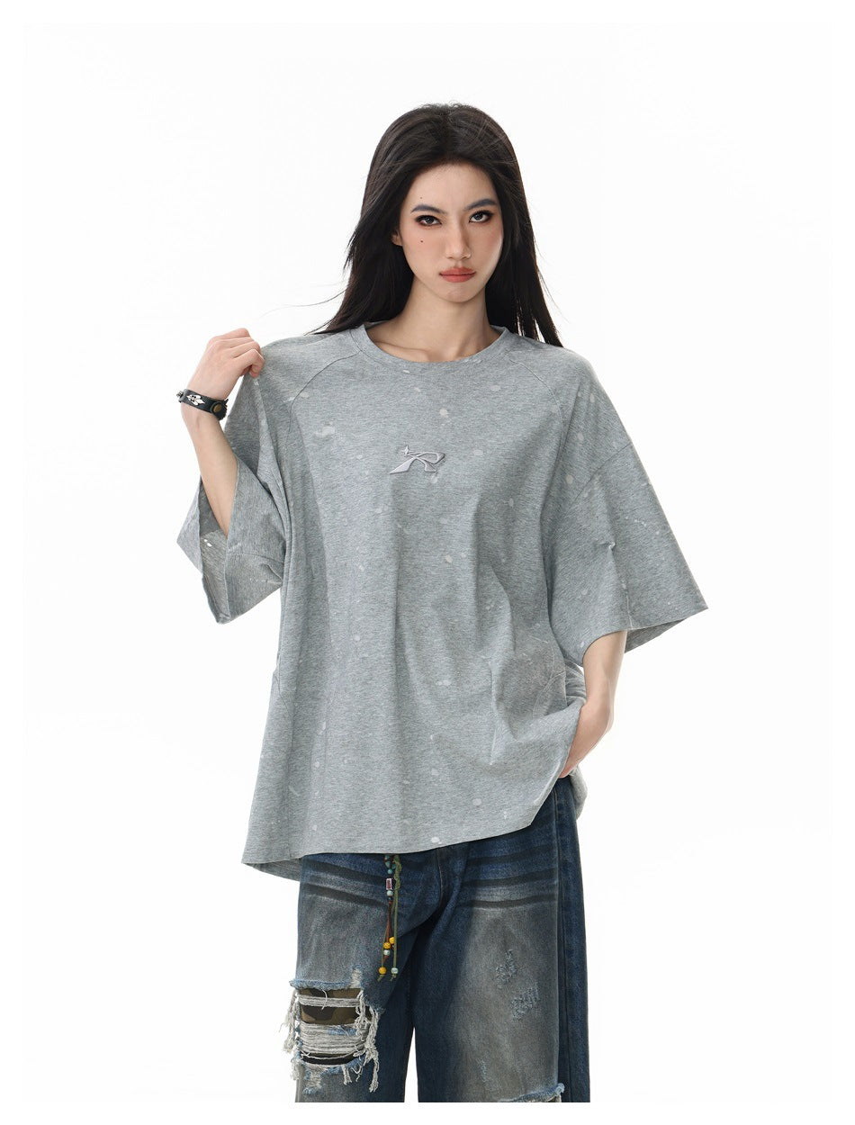 Model wearing a loose light-gray T-shirt with scattered pale marks and a small chest emblem.