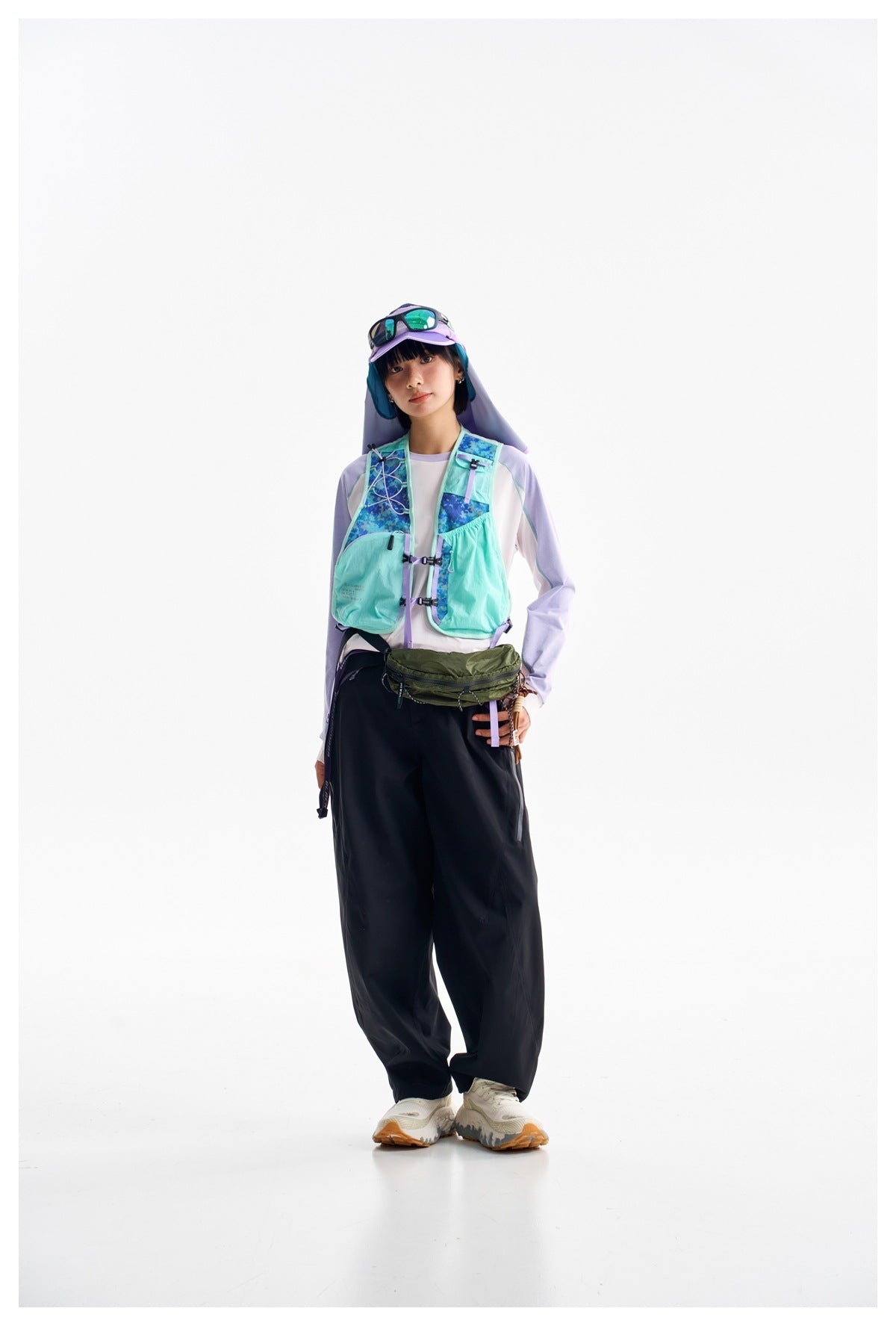 Full-length model wearing a mint hiking vest with blue patterned panels, a lavender top and black trousers.
