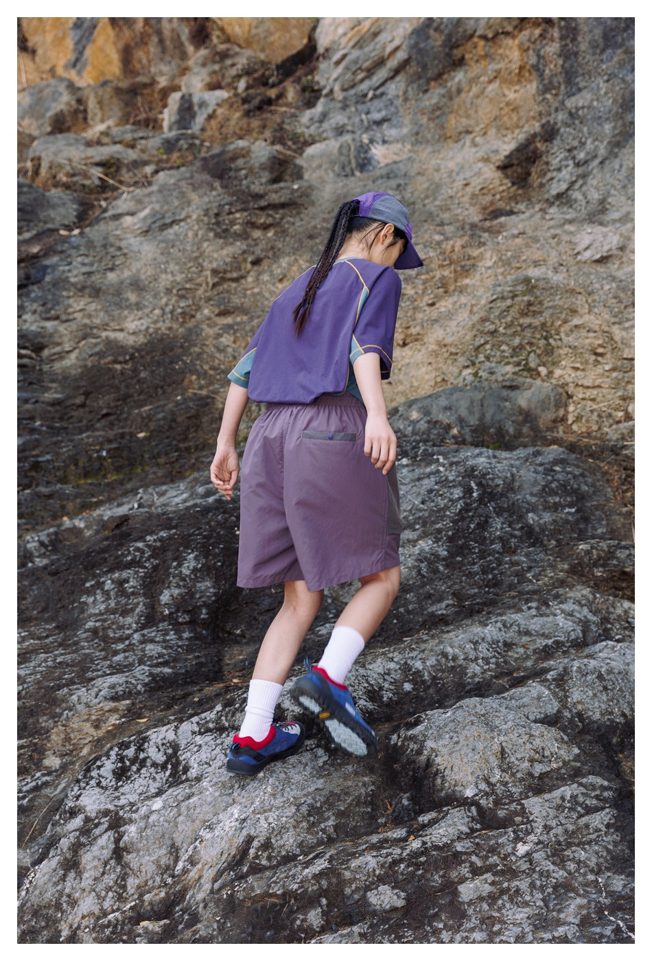 Rear view of a woman wearing a purple color-block T-shirt, lilac shorts and a matching cap while walking on rocks.