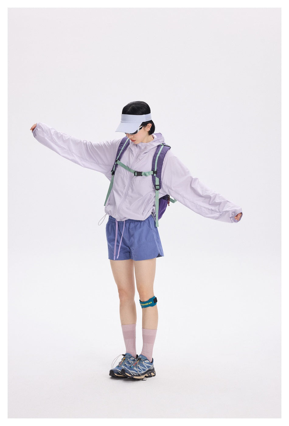 Model wearing a lavender hooded jacket, blue shorts, and a backpack, with arms outstretched.