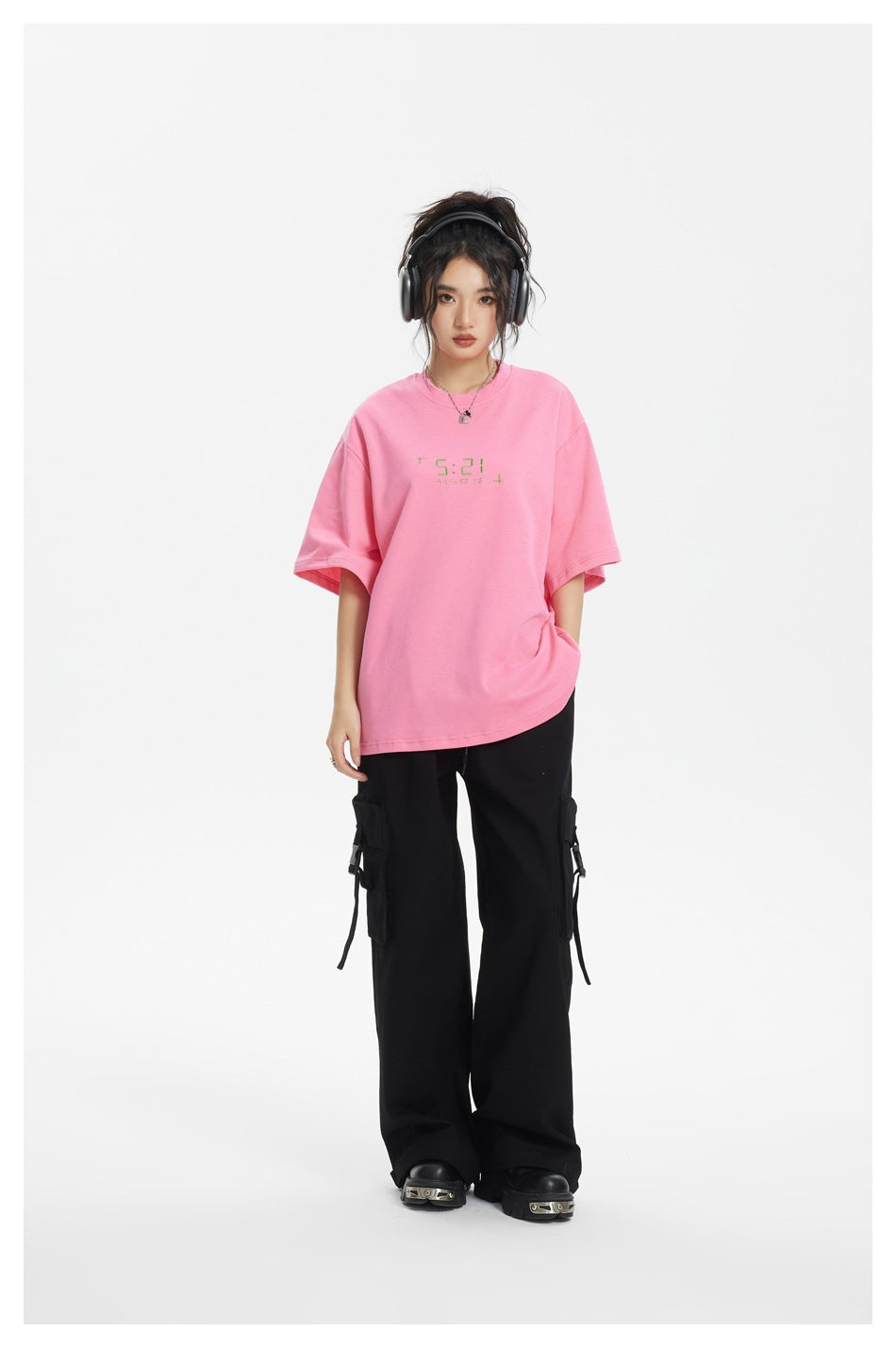 Model wearing an oversized pink 5:21 T-shirt with black cargo pants and headphones, standing front view