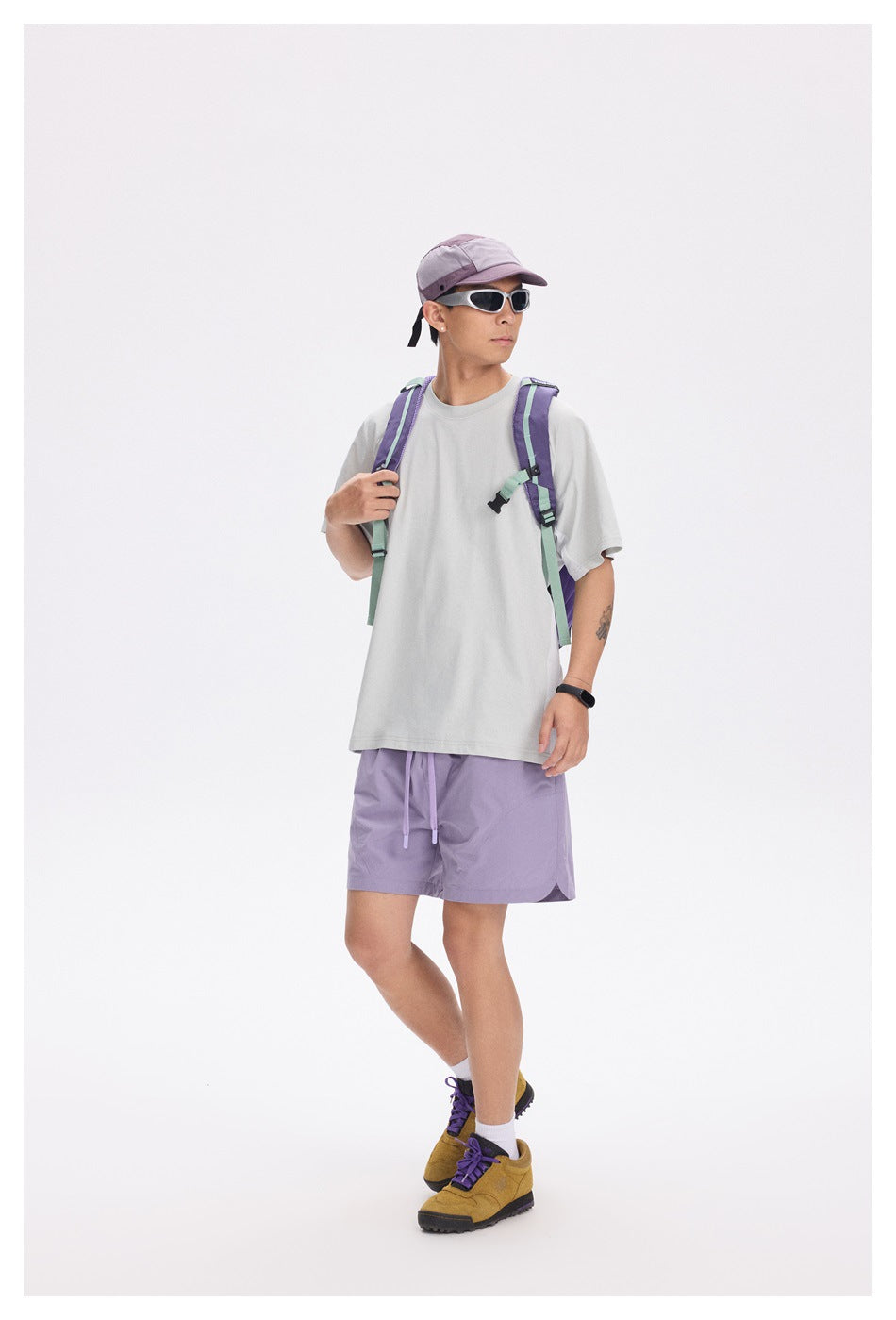 Model in a light gray short-sleeve T-shirt with purple shorts, cap, sunglasses and backpack, full-length view