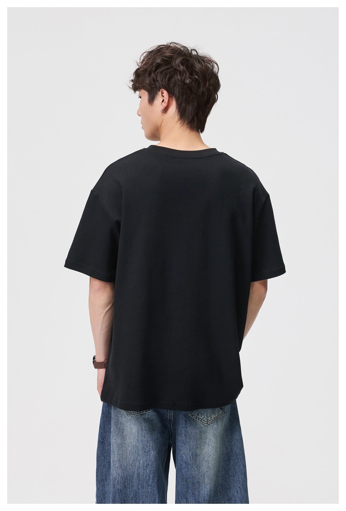 Back view of model wearing a black short-sleeve crewneck T-shirt with dropped shoulders and blue jeans