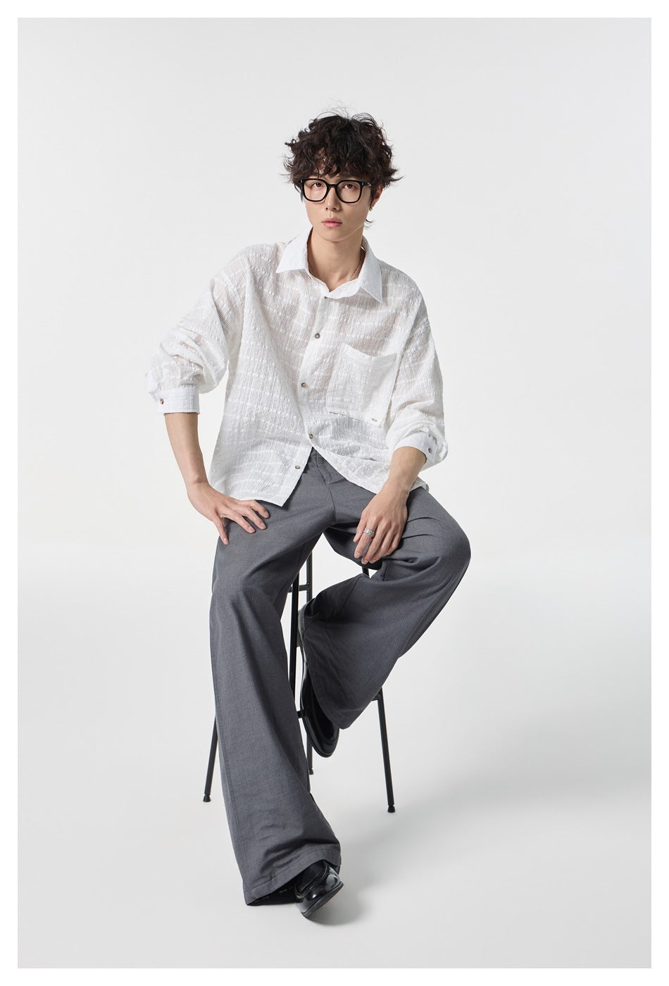 Seated model facing forward in a white textured button-up shirt and gray wide-leg trousers.