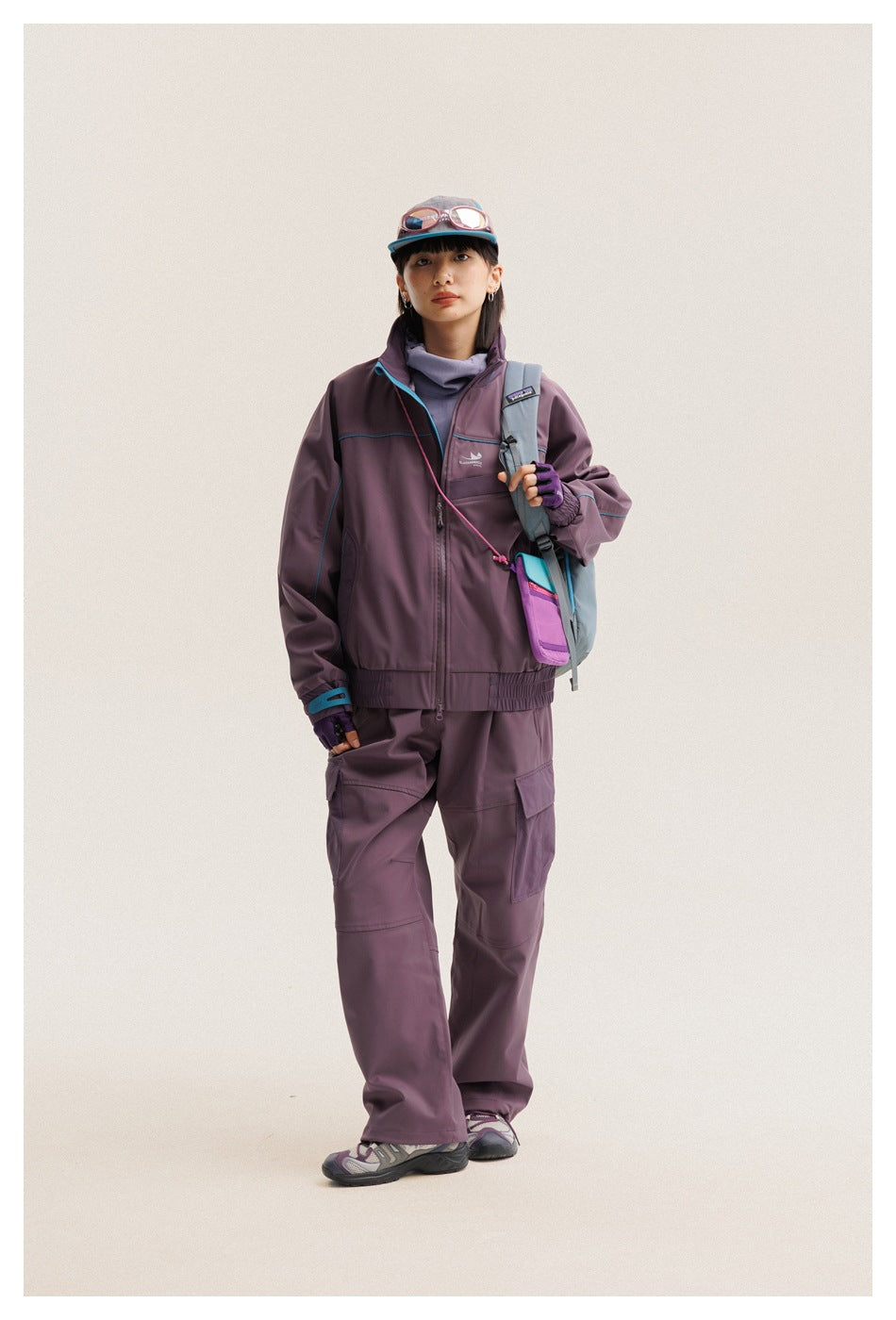 Full-length model wearing a plum zip jacket and cargo pants with turquoise accents, cap, sunglasses, and a shoulder bag.