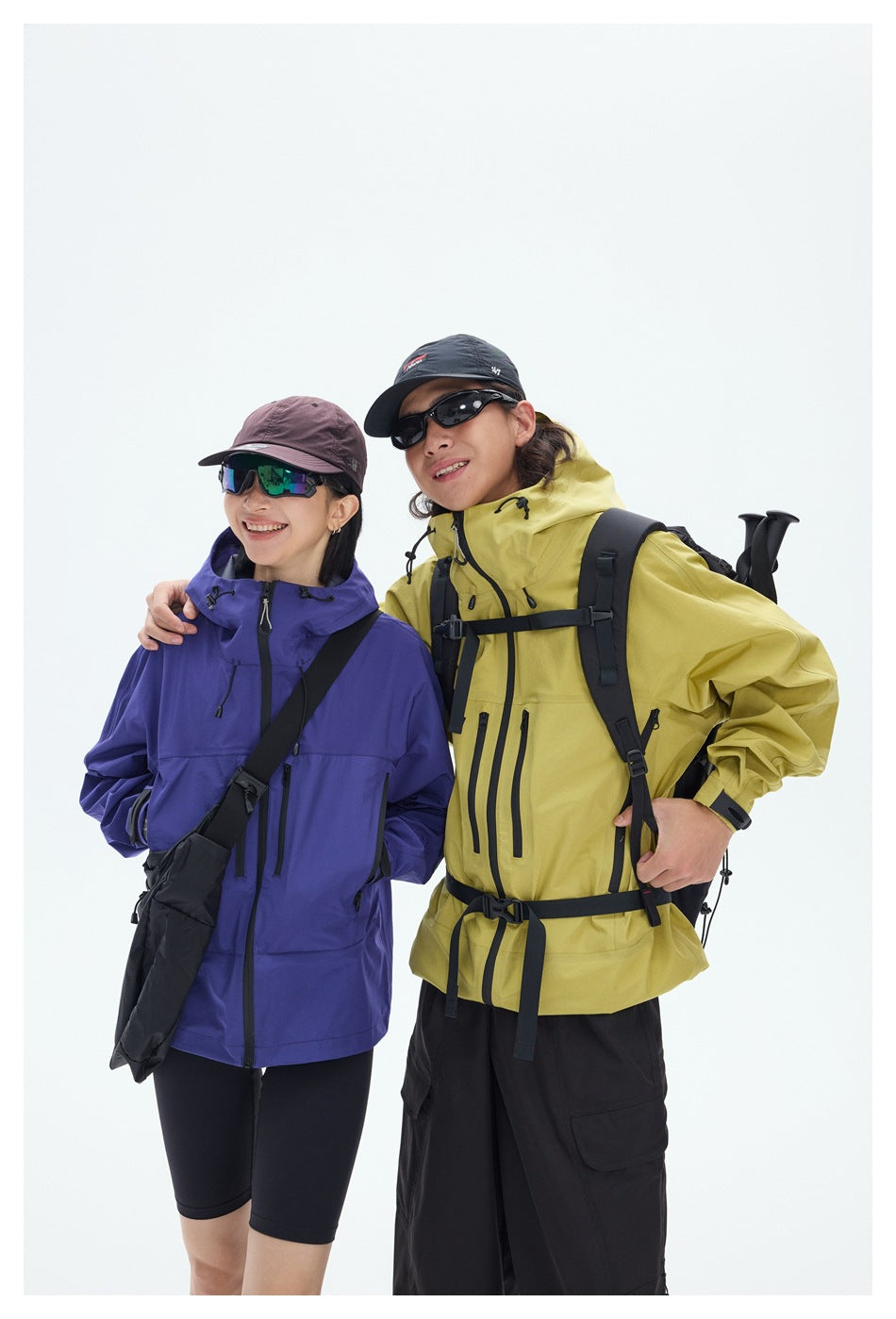 Two models wearing hooded outdoor jackets, one purple and one yellow, with black hiking gear and crossbody bags.