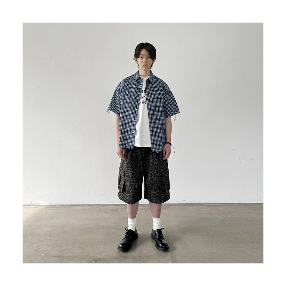 A model wears a blue plaid short-sleeve button-up over a white T-shirt with dark patterned shorts.