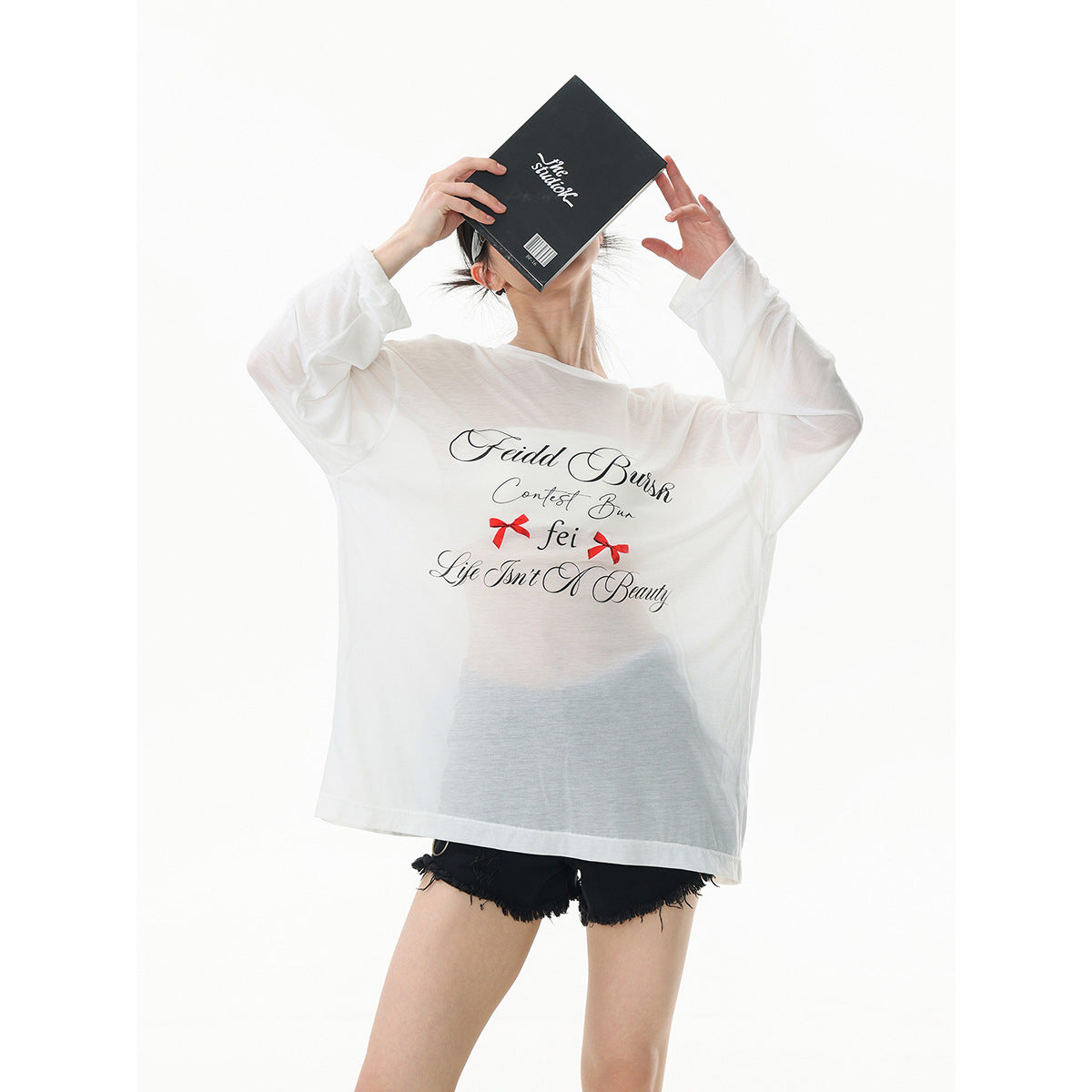 Model wearing an oversized sheer white graphic tee with black script and red bows, holding a black book.