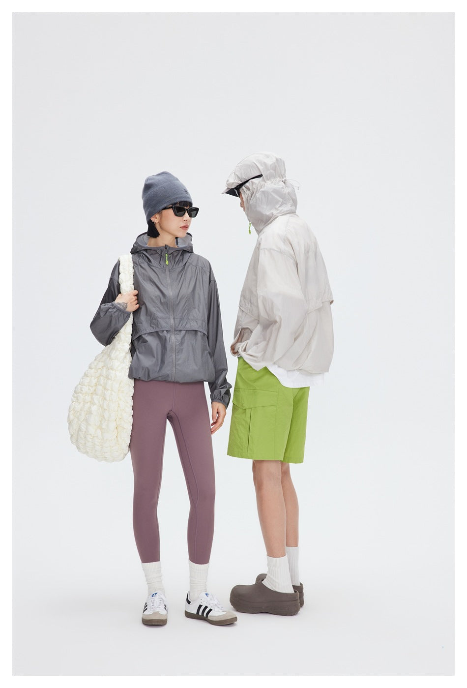 Two models wear hooded jackets in pale beige and charcoal gray, with contrasting outfits and accessories.