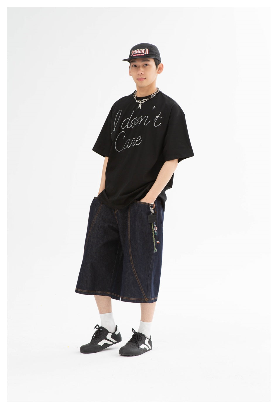 Full-length model wearing a black oversized rhinestone-script T-shirt, dark denim shorts, and a black cap.