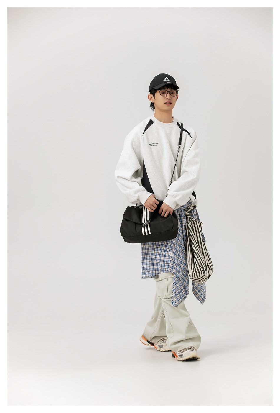 Full-length model wearing a light gray sweatshirt with black curved panels and layered casual accessories.