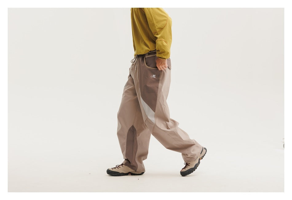 Model wearing loose taupe trousers with darker panels and a pale curved side stripe, shown in profile while walking.