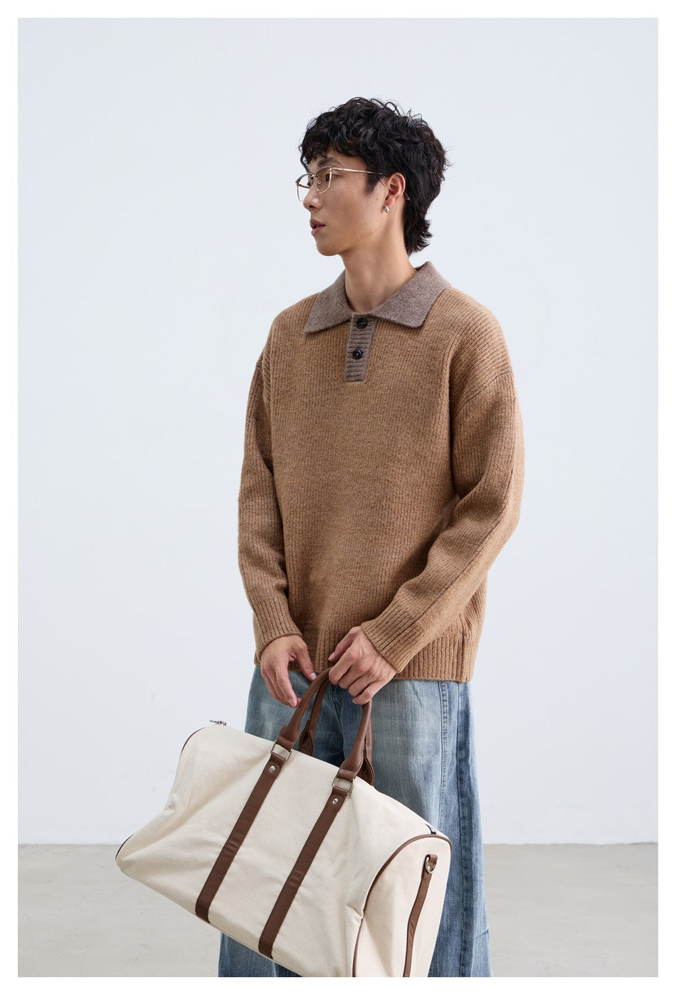 Model wearing a camel-brown knit polo sweater with a taupe collar, holding a cream duffel bag.
