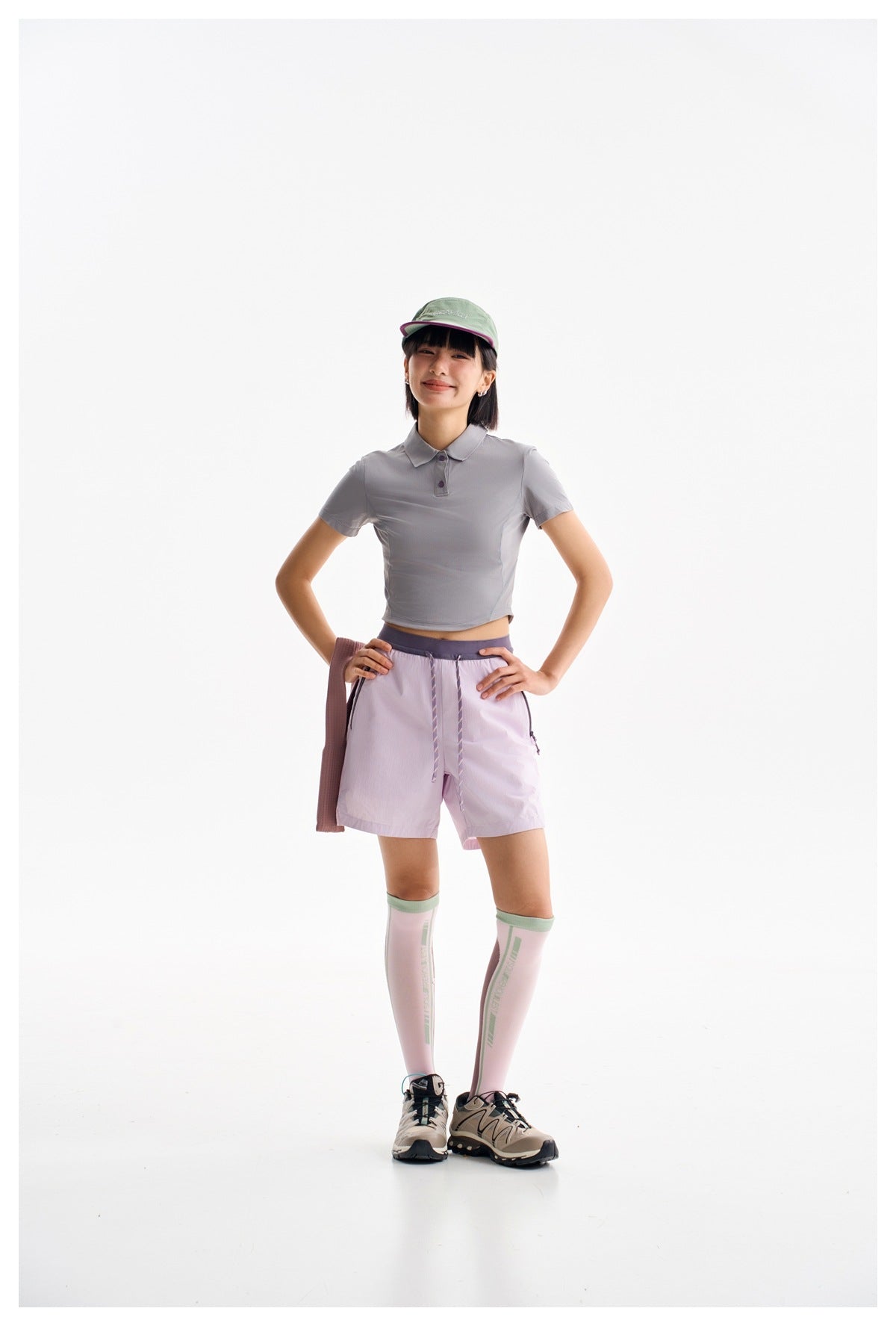 Front view of a model wearing a gray cropped polo, lilac shorts, mint cap and pink knee-high socks.