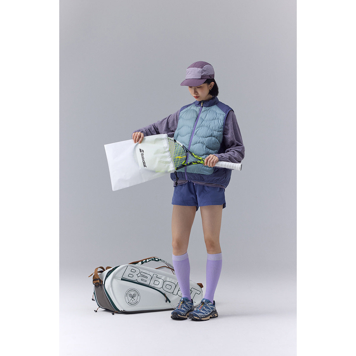 Model wearing a pale blue quilted vest with purple trim and holding a tennis racket beside a racket bag.