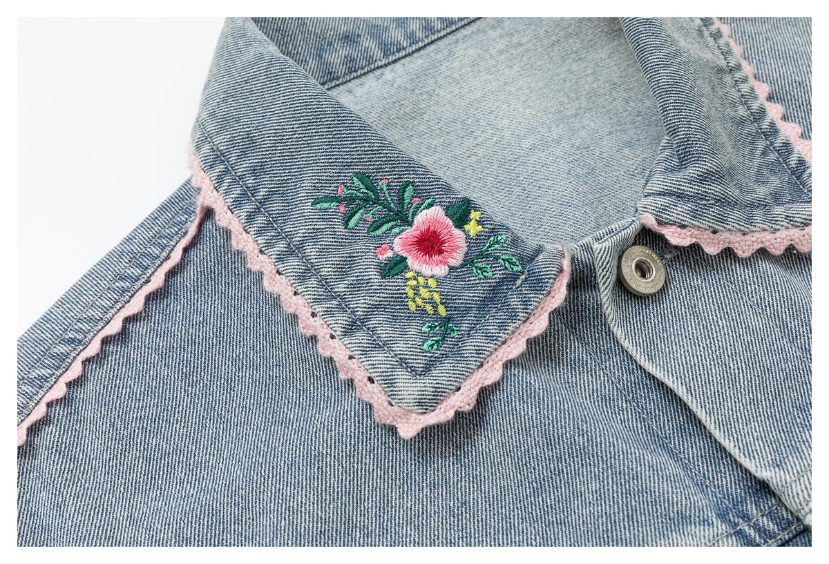 Close-up of a blue denim jacket collar with a pink floral embroidered motif, scalloped trim, and metal button.
