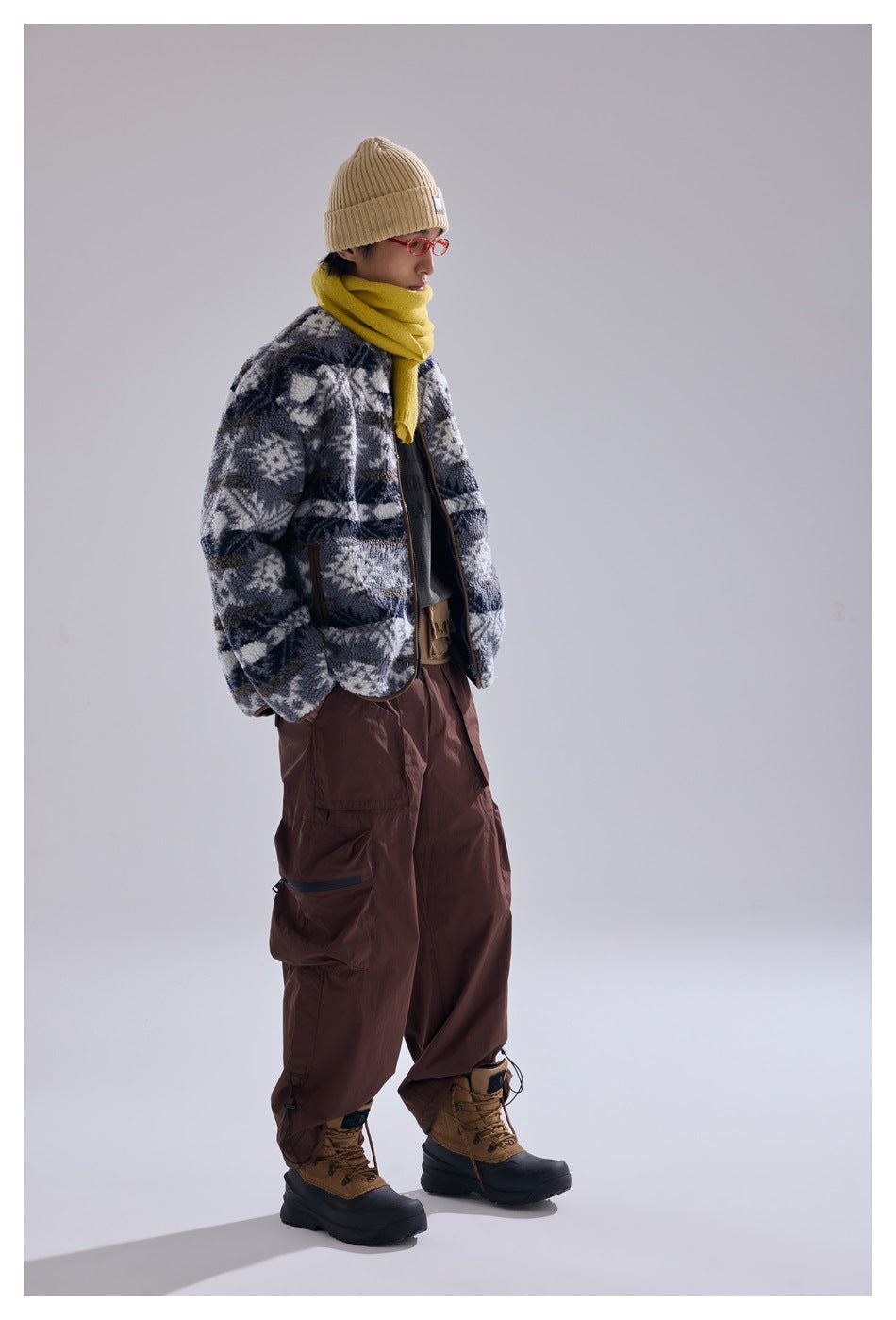 Model in gray geometric pattern fleece-like jacket with hands in pockets, yellow scarf and brown cargo pants, side view