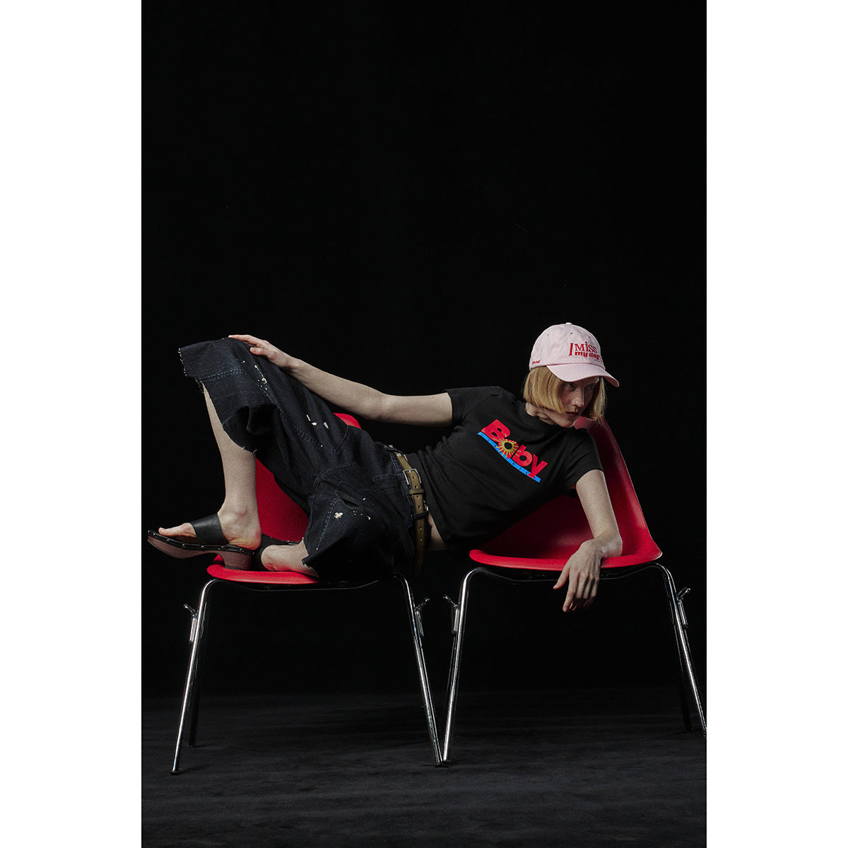 Model reclining across red chairs, wearing a black graphic T-shirt and dark wide-leg jeans.