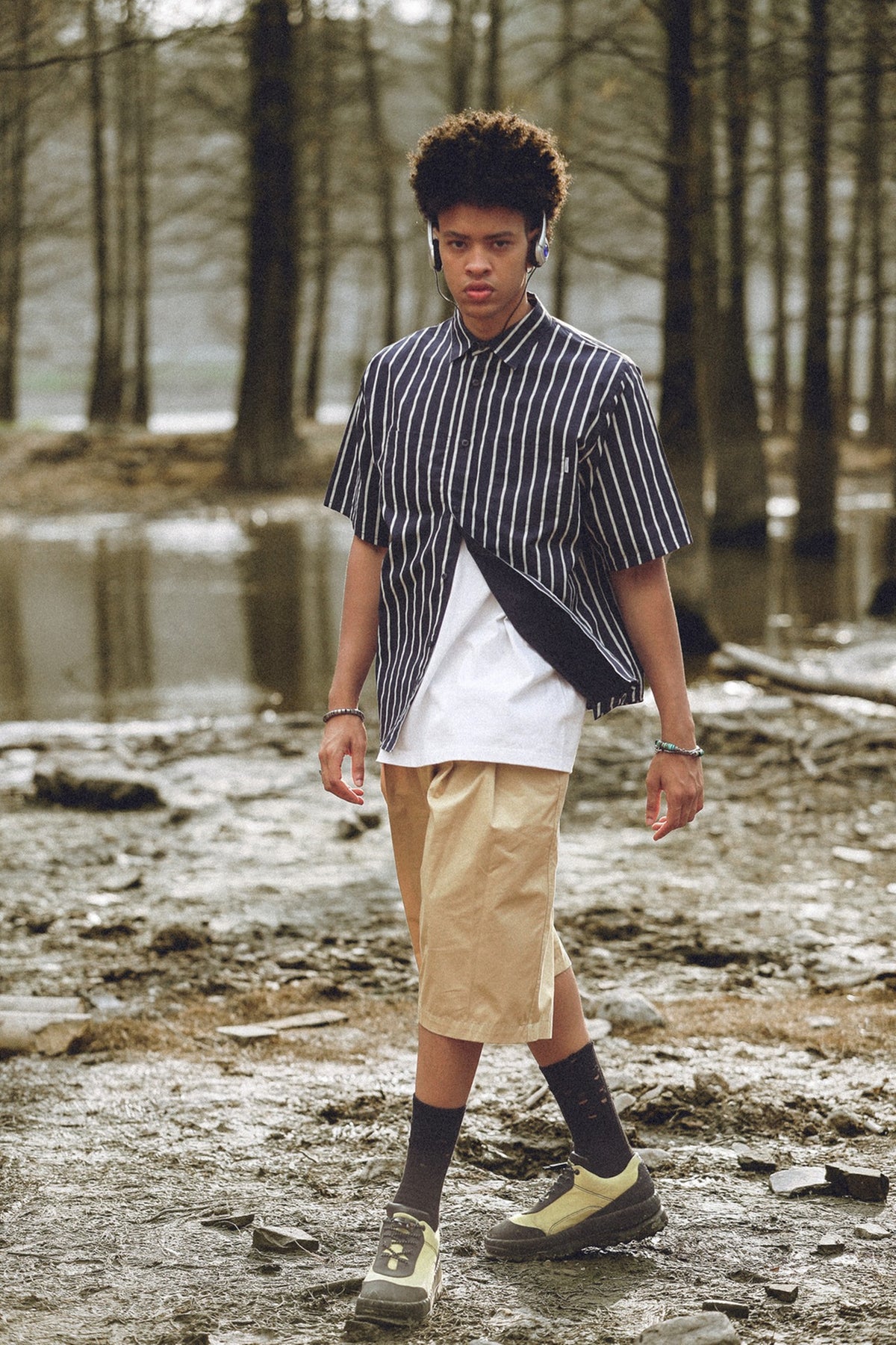 Model outdoors wearing a black-and-white striped short-sleeve shirt over a white tee and tan knee-length shorts.