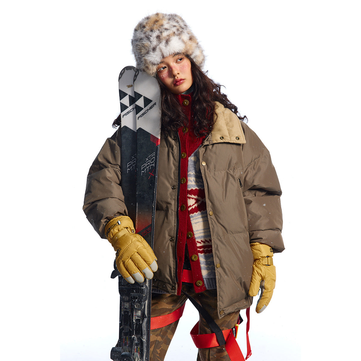 Model wearing a taupe puffer jacket over a red patterned cardigan, holding skis against a white background.