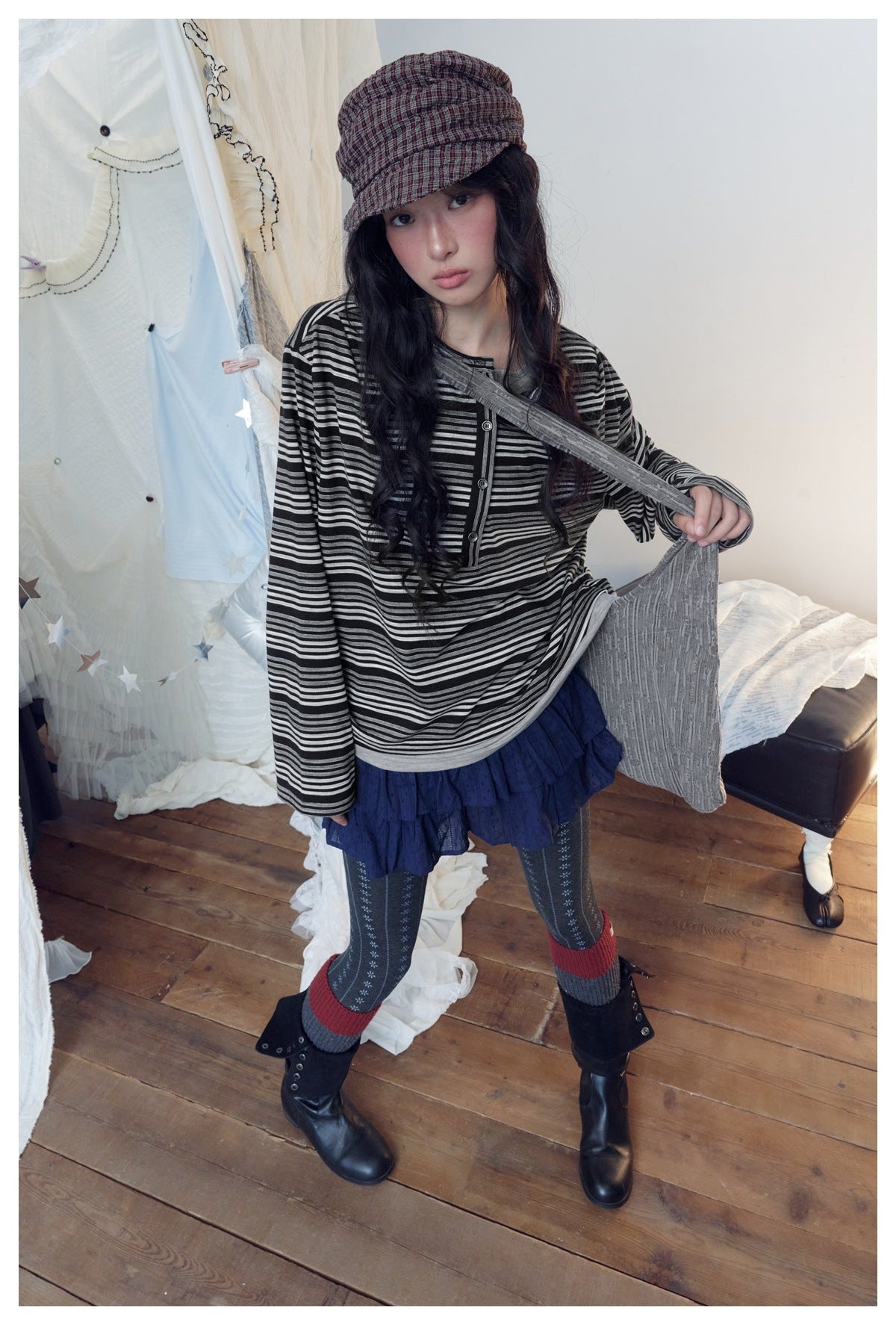 Model wearing a black and white striped henley top with a navy ruffle skirt, patterned tights, boots and a plaid cap