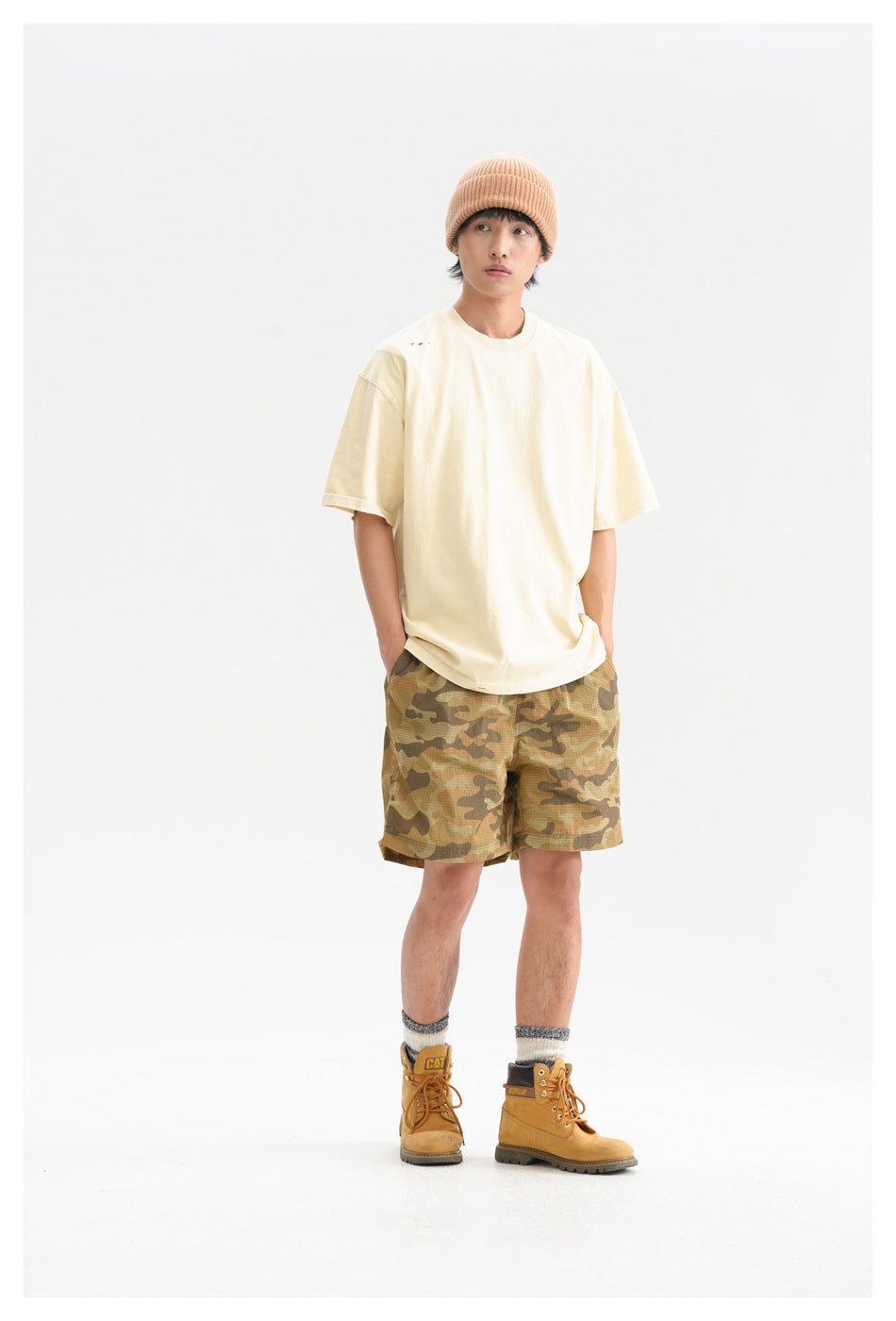 Model in a light yellow oversized T-shirt, camo shorts, beanie and tan boots, hands in pockets