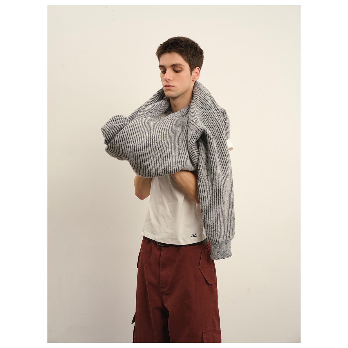 Model holding up a light gray ribbed crewneck sweater, revealing a white T-shirt and rust-red cargo pants.