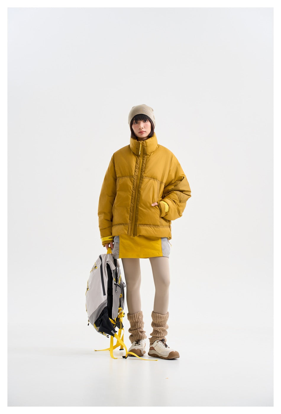 Model wearing a mustard-yellow padded jacket with a high collar, shown from the front with a backpack.