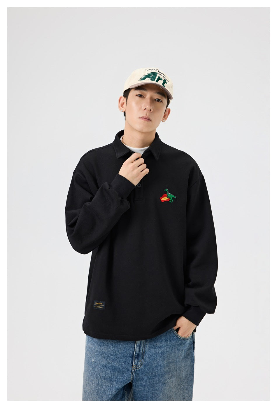 Model in black polo-collar top with chest dinosaur embroidery, hand at collar, wearing cap and jeans