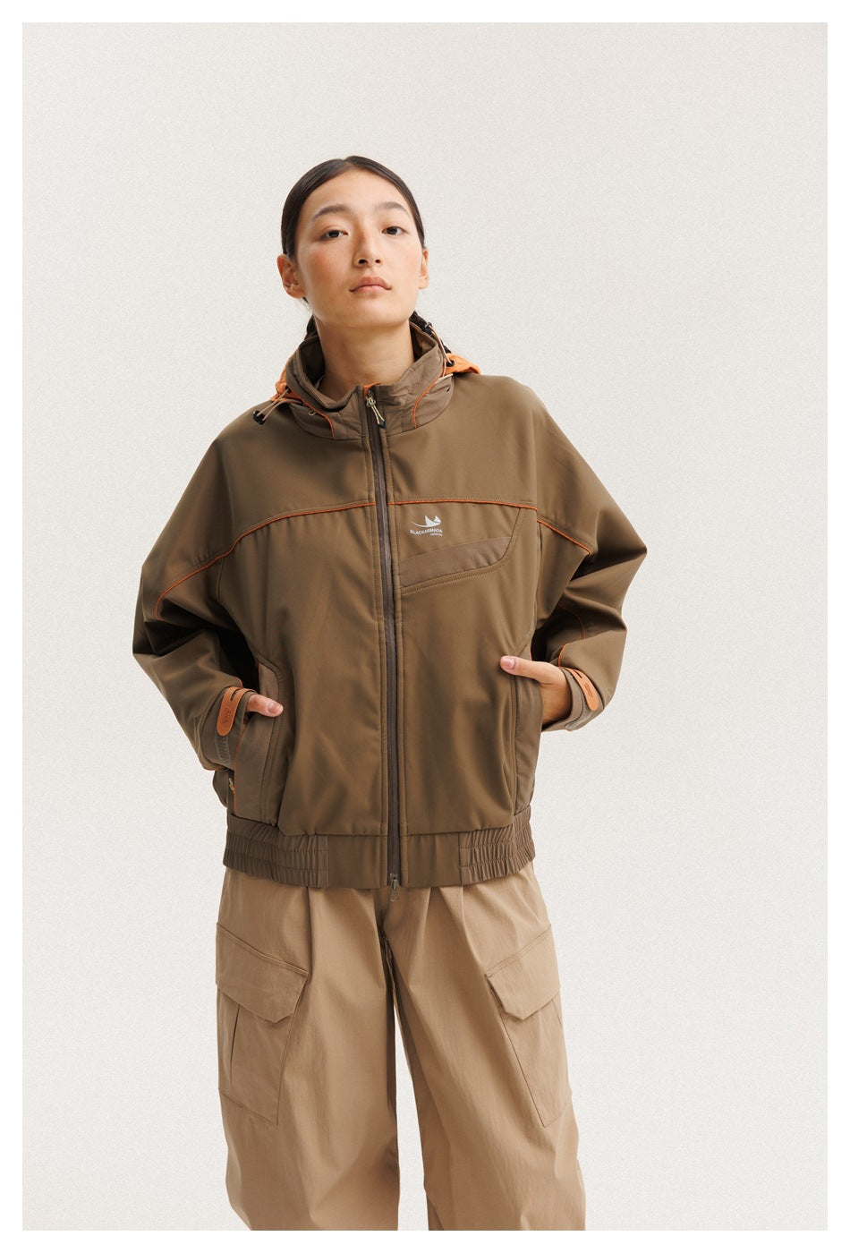 Model wearing a brown hooded jacket with orange trim and tan cargo pants, shown from the front with hands in pockets.