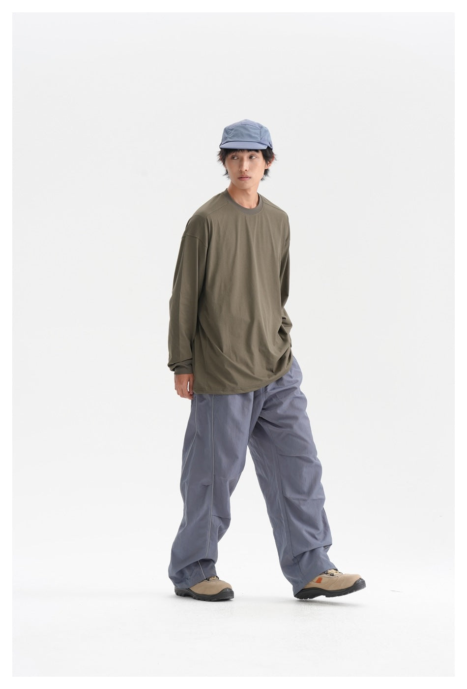 Model walking in an army green long-sleeve top with gray wide-leg pants, blue cap and beige shoes, glancing sideways