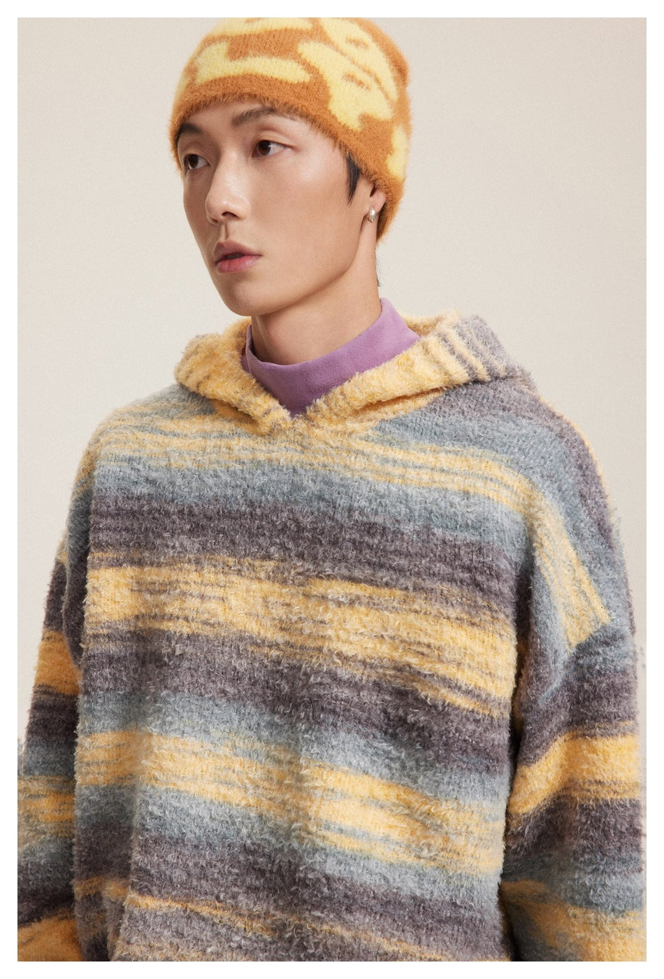 Close view of a model wearing a gray, yellow, and brown striped hooded sweater and an orange-yellow beanie.
