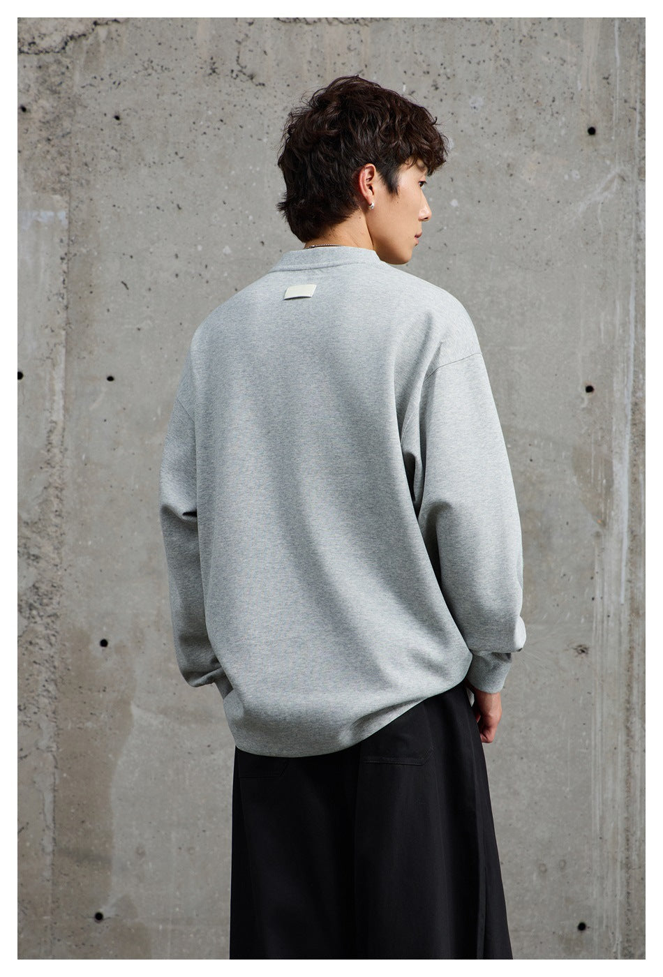Rear view of a model wearing an oversized light gray crewneck sweatshirt with a small blank patch at the back neck.
