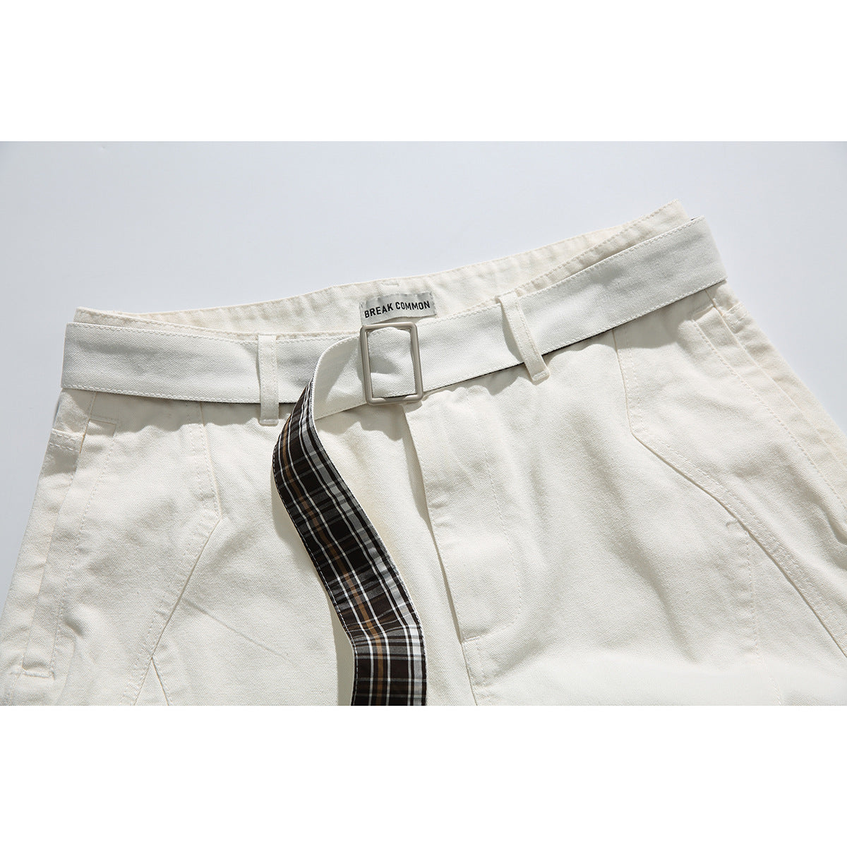 Close-up of ivory shorts with a square buckle belt and a dark brown plaid belt end.