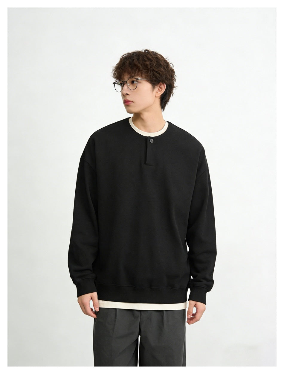 Man wearing black button-placket sweatshirt over a cream tee with gray trousers, three-quarter front view