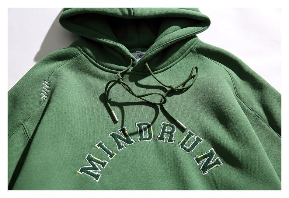 Close-up of a green hoodie with drawstrings, white zigzag stitching, and embroidered arched lettering.