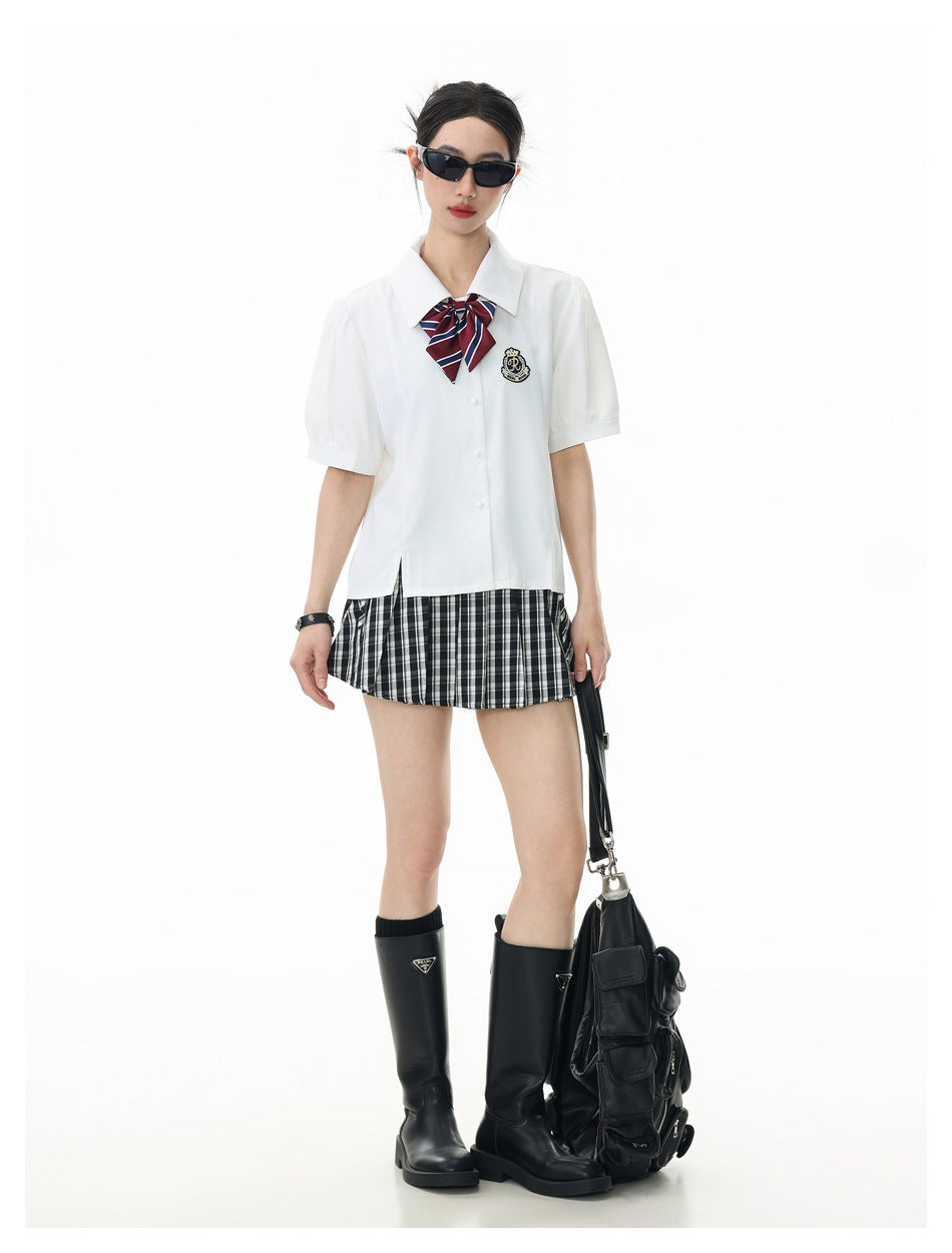 Model wearing a white short-sleeve shirt with a striped bow and crest patch over a black and white pleated plaid skirt