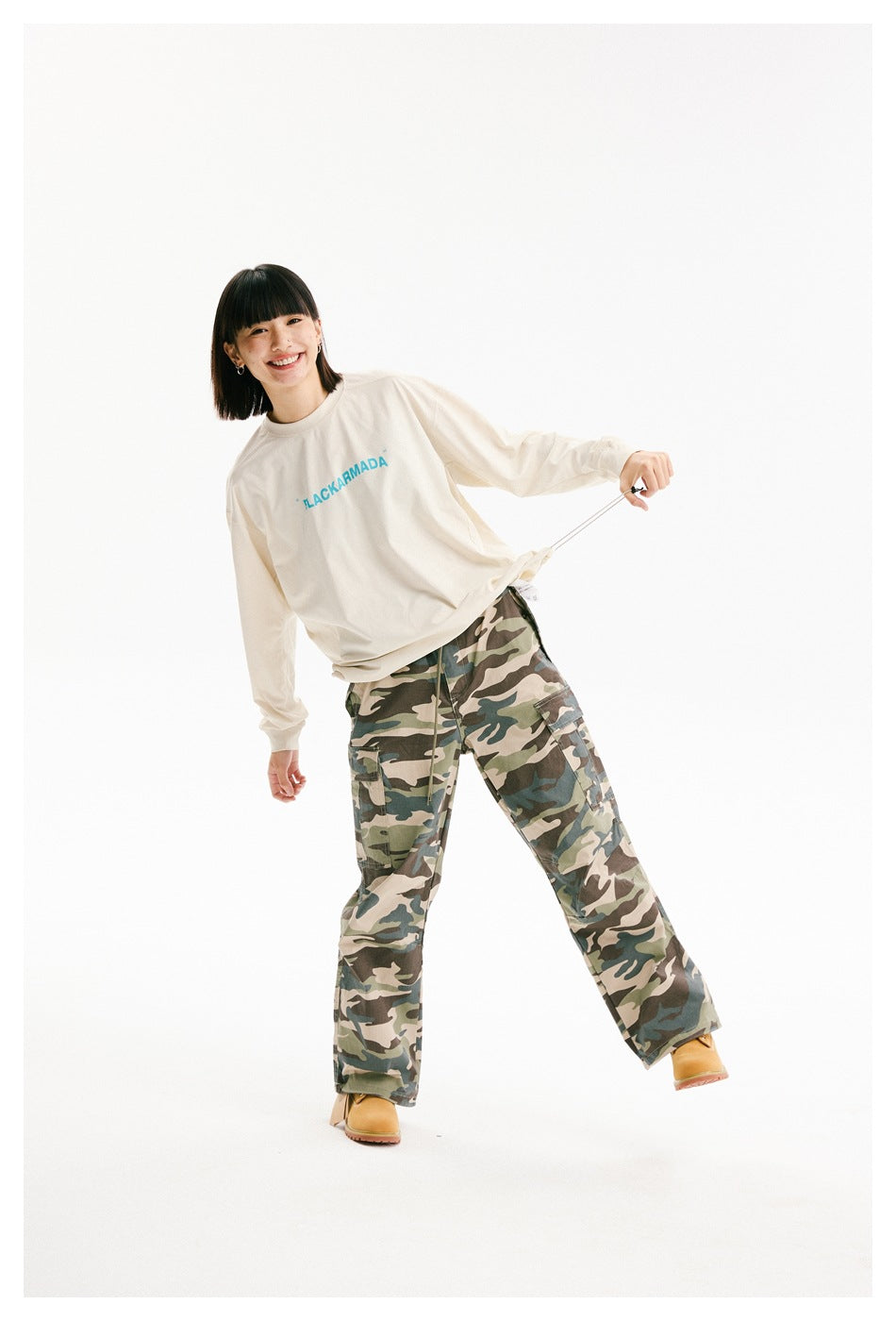 Model wearing an ivory long-sleeve shirt and camouflage cargo pants, pulling the shirt hem outward to show its fit.
