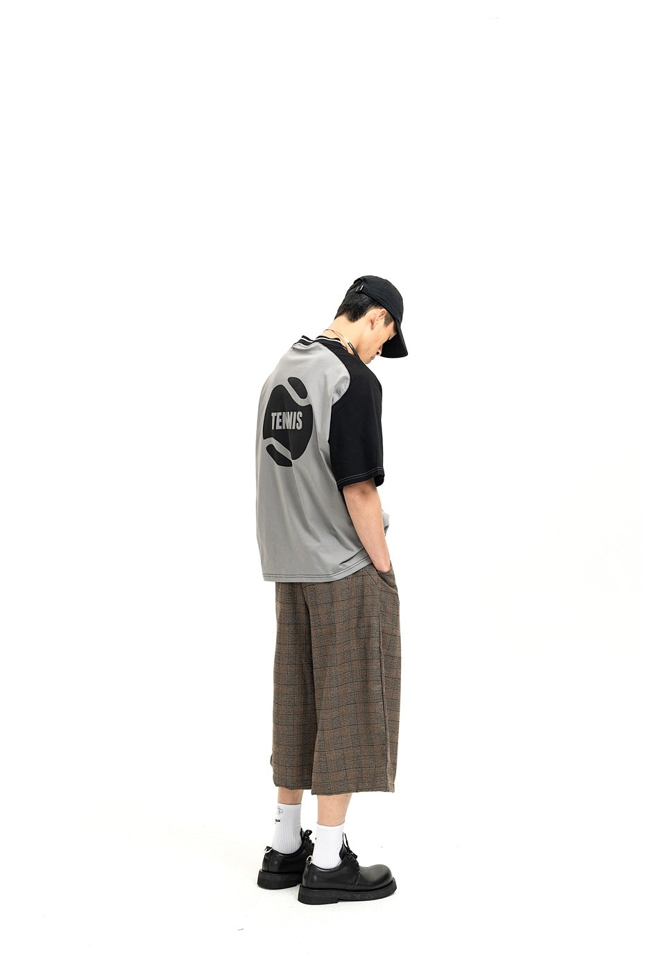 Back view of man wearing gray and black raglan T-shirt with tennis ball graphic and plaid cropped shorts