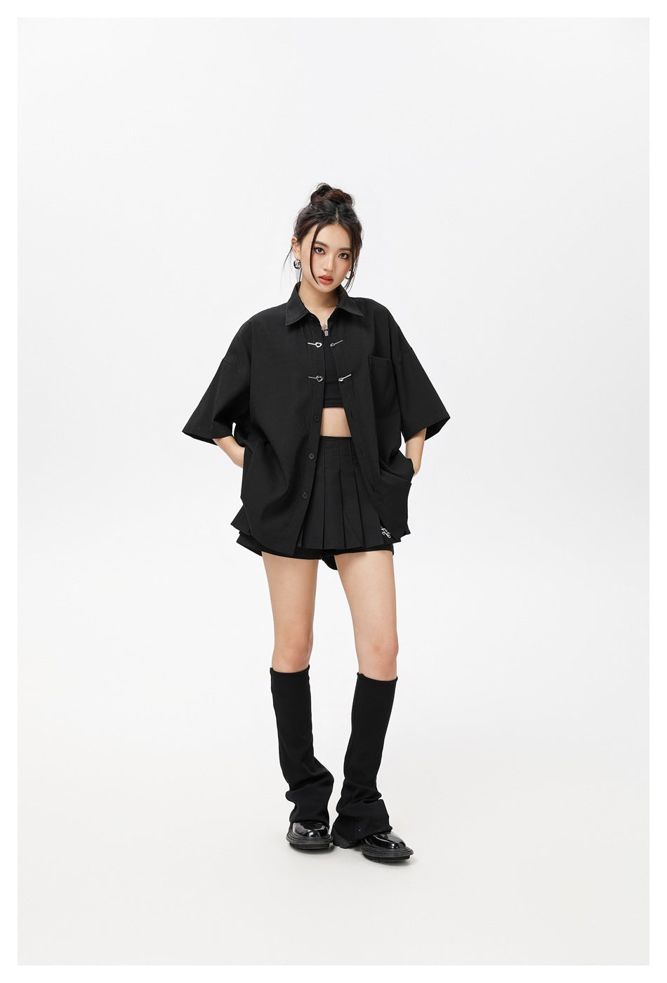 Model wearing the black oversized short-sleeve shirt open with hands in pockets, over a crop top and pleated skirt