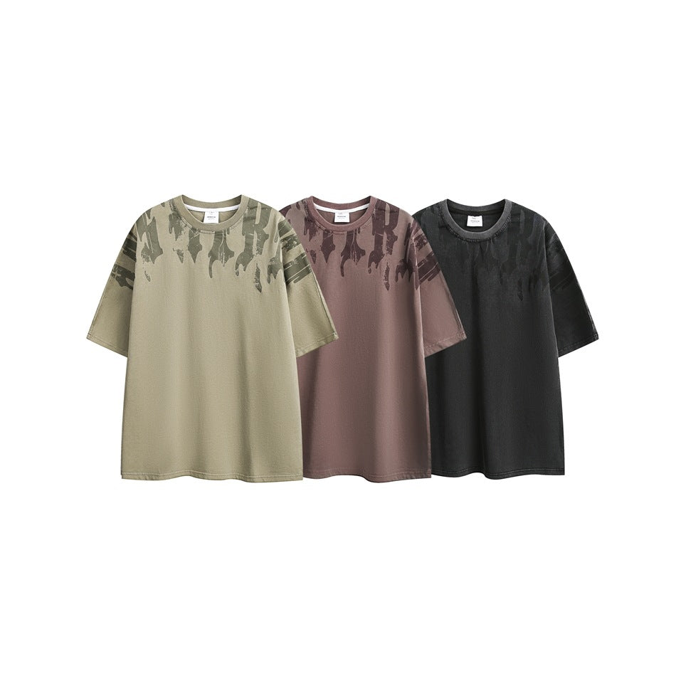 Three short-sleeve T-shirts shown together in olive, mauve, and charcoal, each with a dark abstract shoulder print.