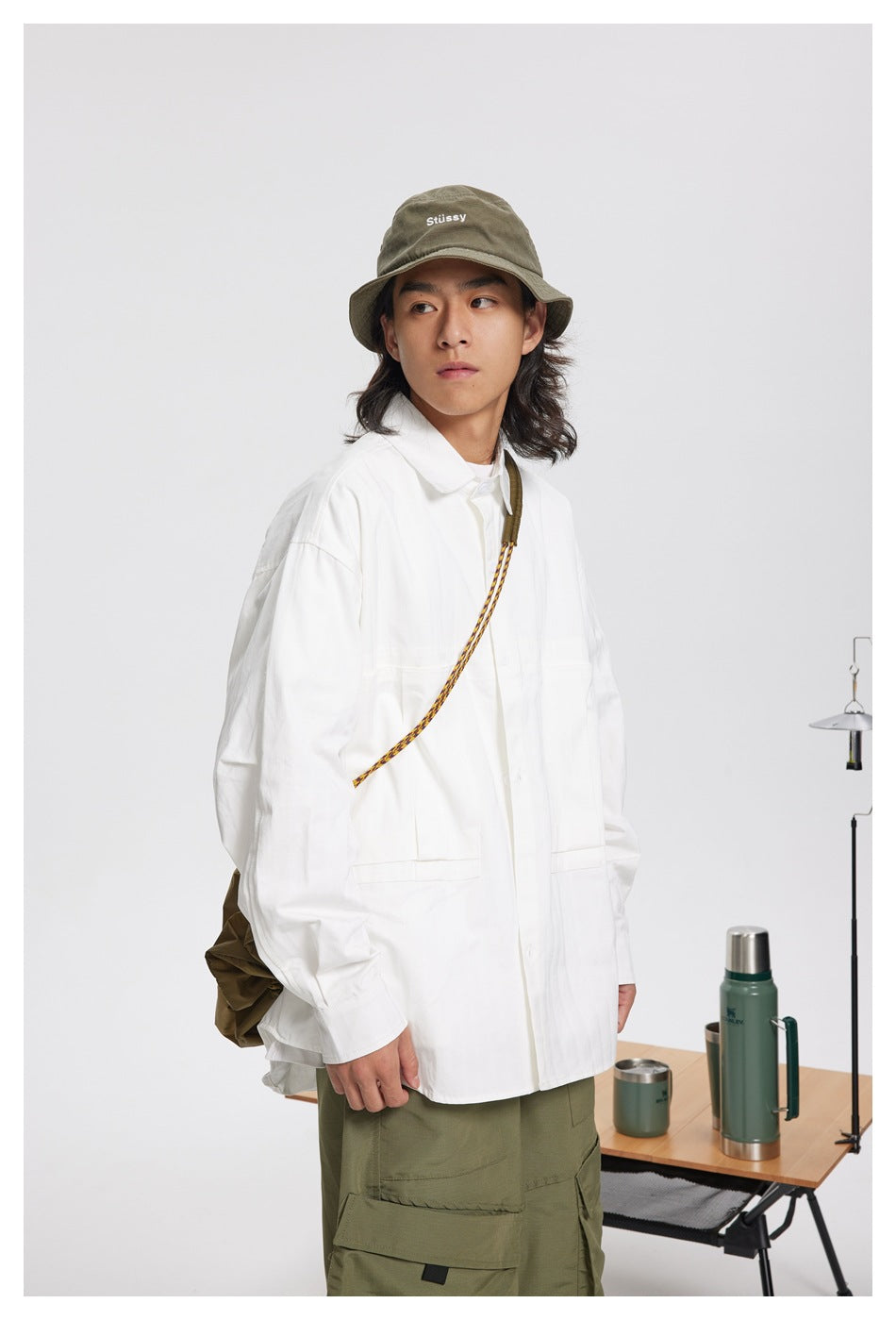 Model wearing a white button-up overshirt, olive cargo pants, and an olive bucket hat with a shoulder bag.