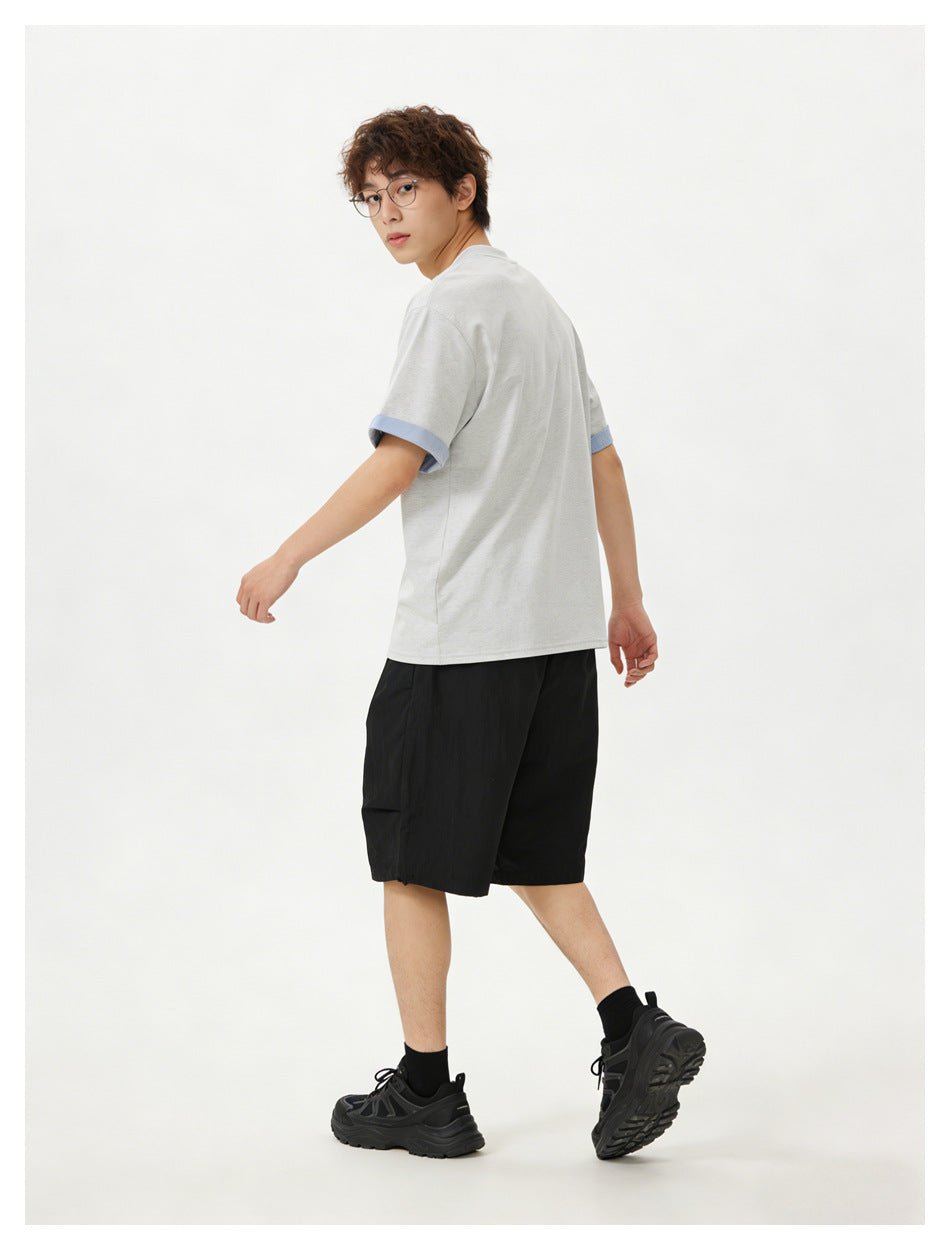 Model walking and looking back, wearing a light gray T-shirt with blue cuffs, black shorts, and black sneakers