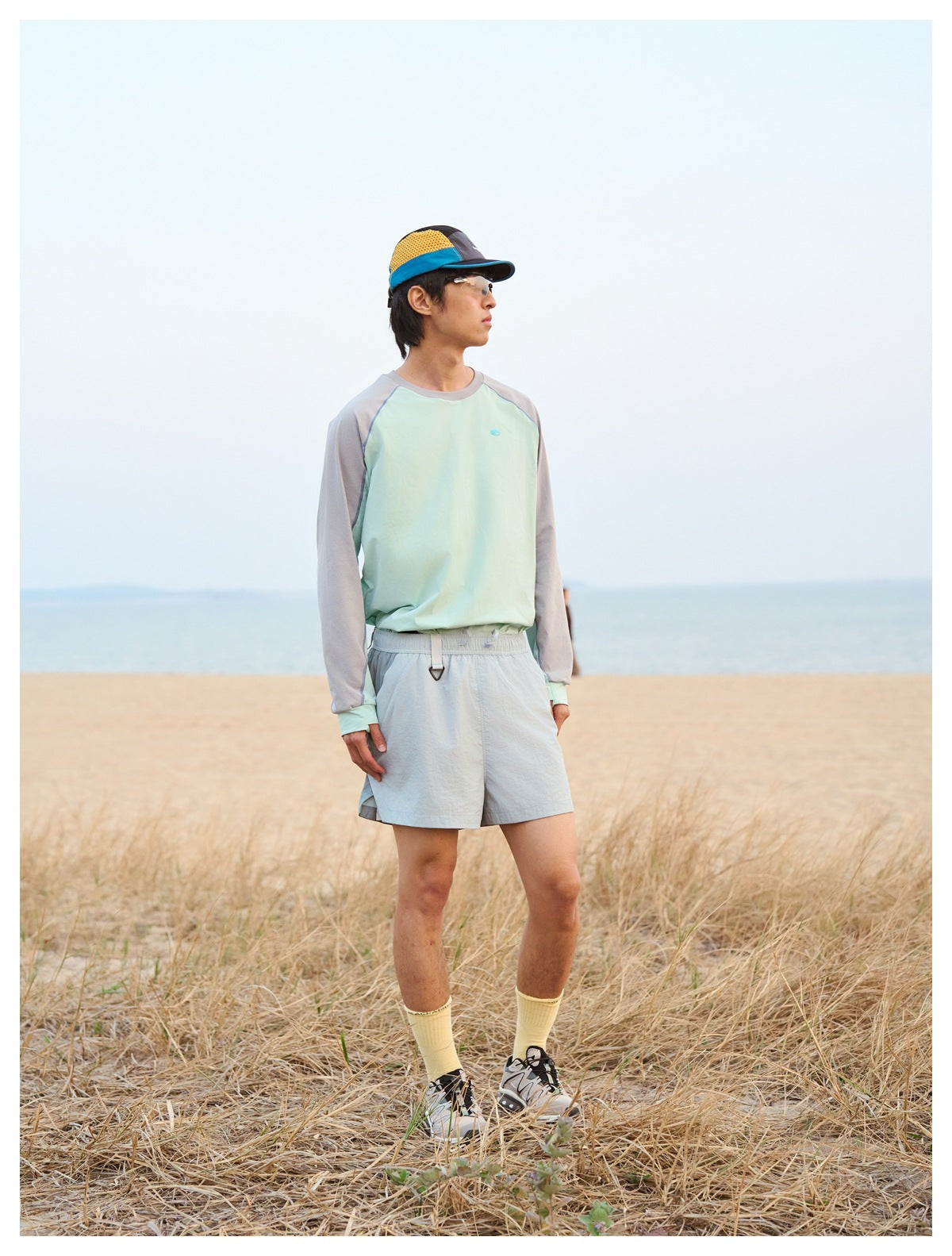 Man wearing a mint and gray raglan long-sleeve top and pale gray shorts by the beach.