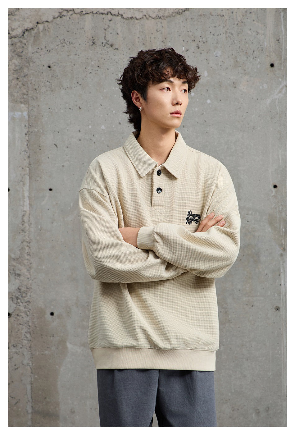 Model with arms crossed wearing a relaxed beige polo-collar sweatshirt and gray trousers against concrete